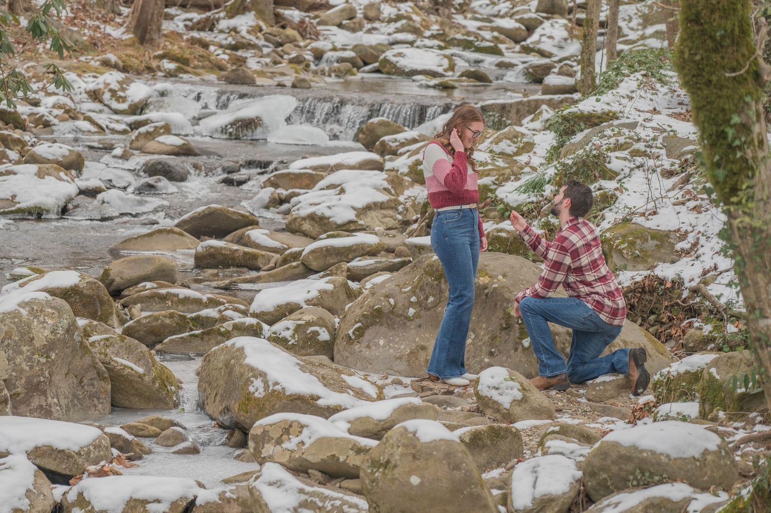 A proposal moment captured on snow-covered rocks with a winter waterfall in the background.