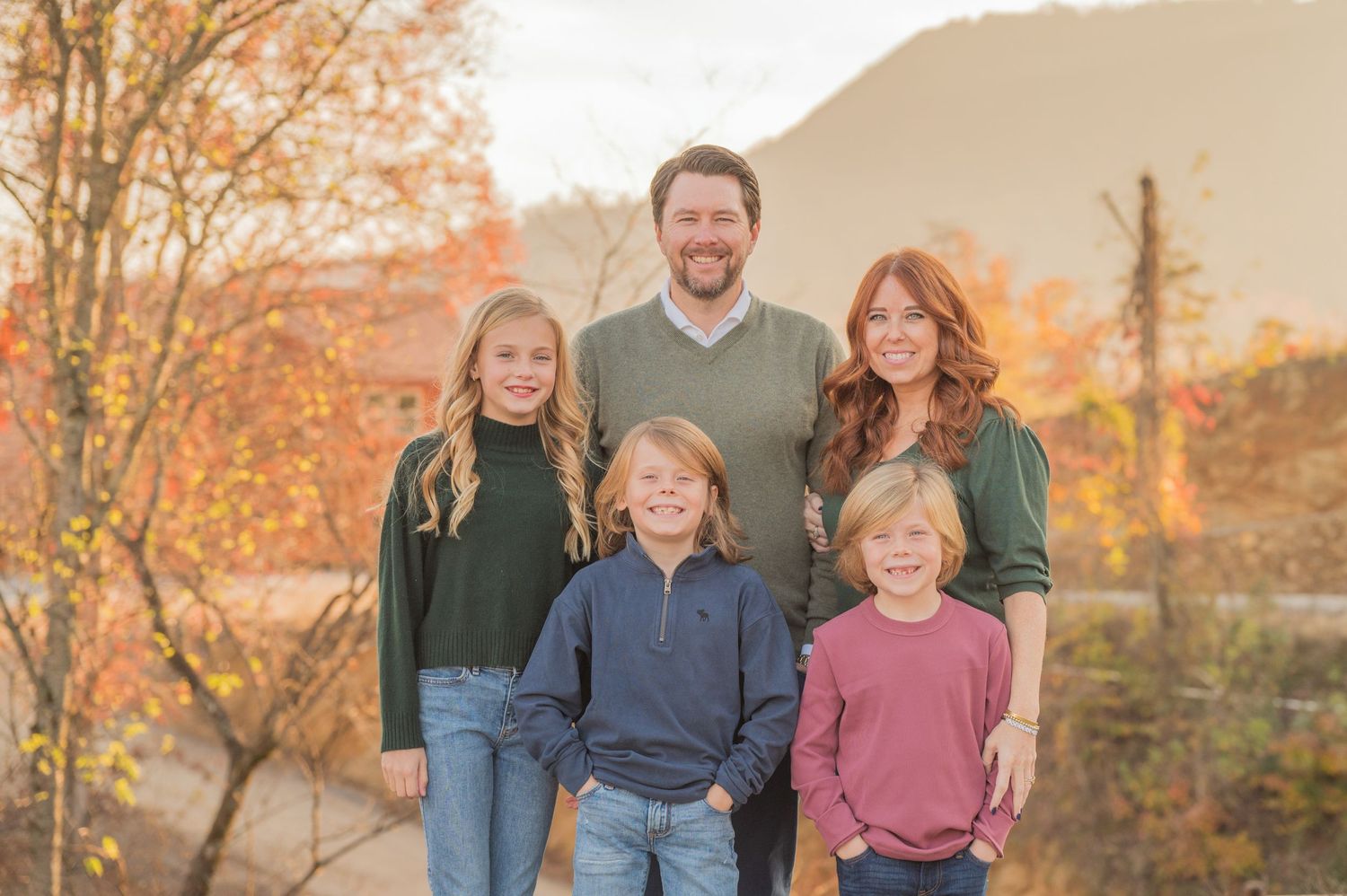 A family of five poses outdoors during autumn with fall foliage and a mountain backdrop at sunset in Sevierville TN.