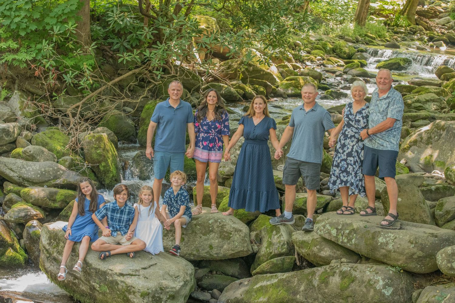 A group of people pose together on large rocks by a peaceful stream in a lush green forest setting by Gatlinburg Photographer.