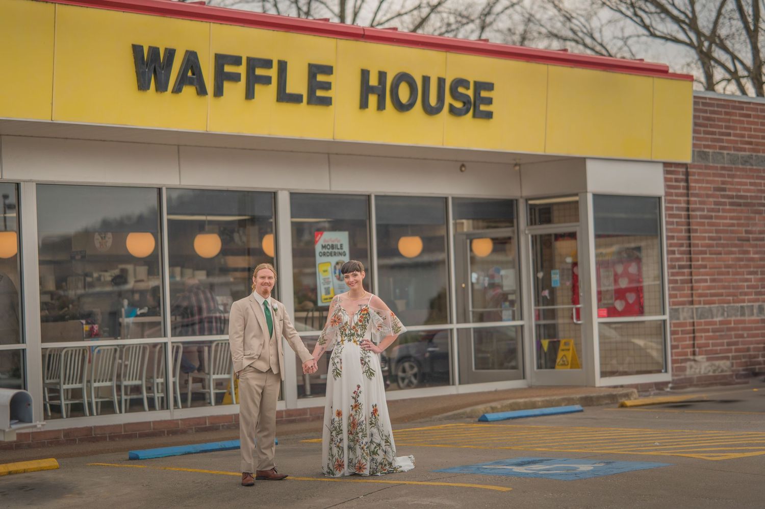Couple stands outside the entrance of a classic Waffle House restaurant in front of yellow signage.