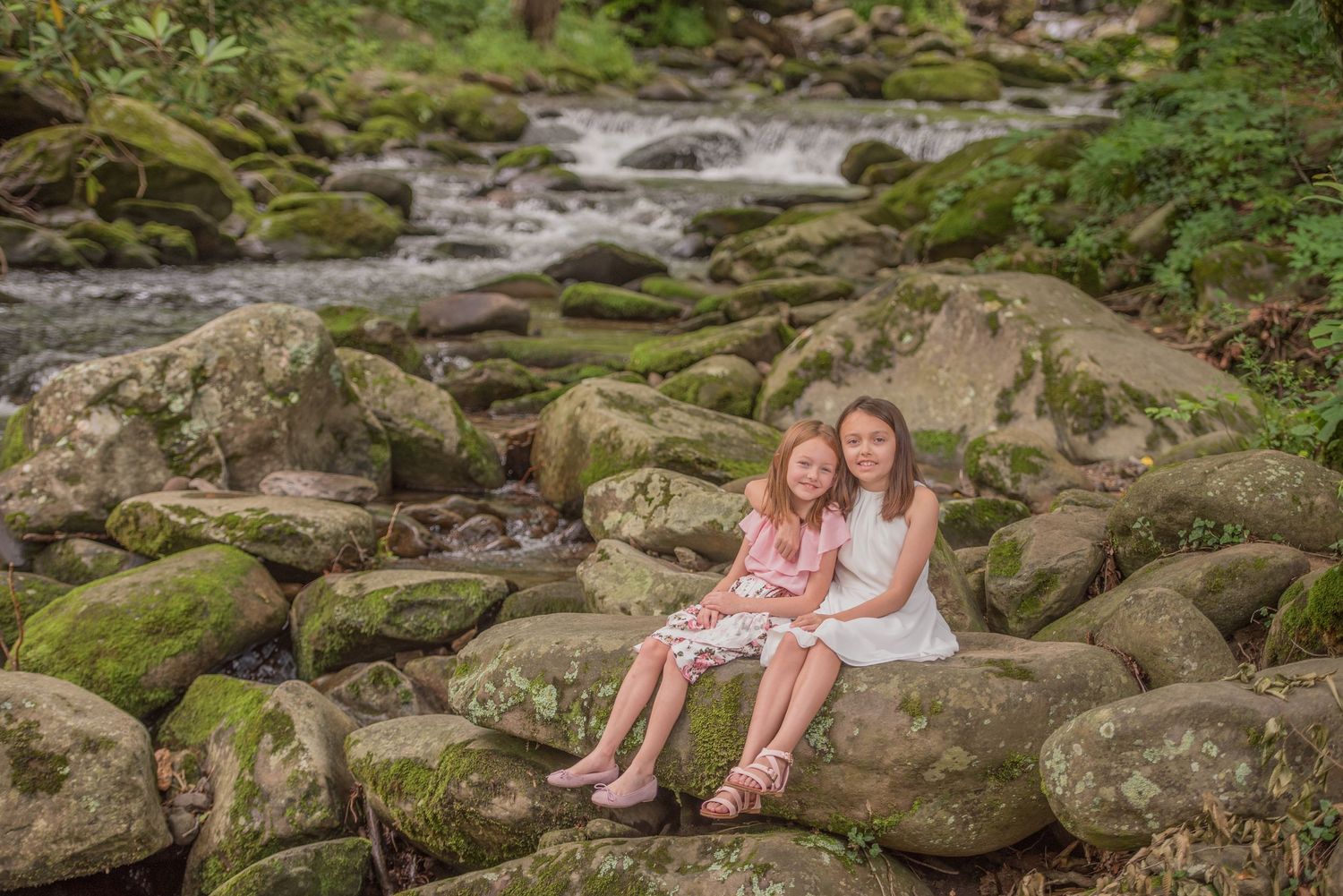 Two people sit together on moss-covered rocks beside a flowing stream in a serene forest setting.
