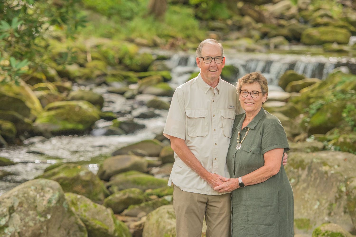 Couple stands together by scenic mountain stream with mossy rocks and flowing water in background by Sevierville Photographer.