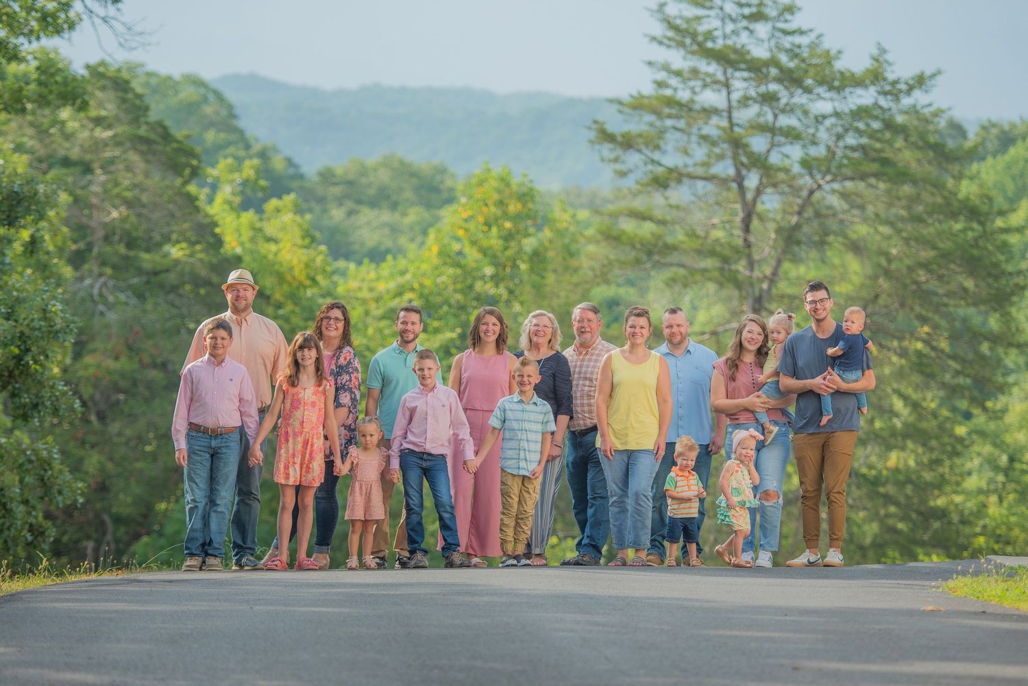 A large group stands together on a scenic mountain road surrounded by lush green trees on a sunny day.