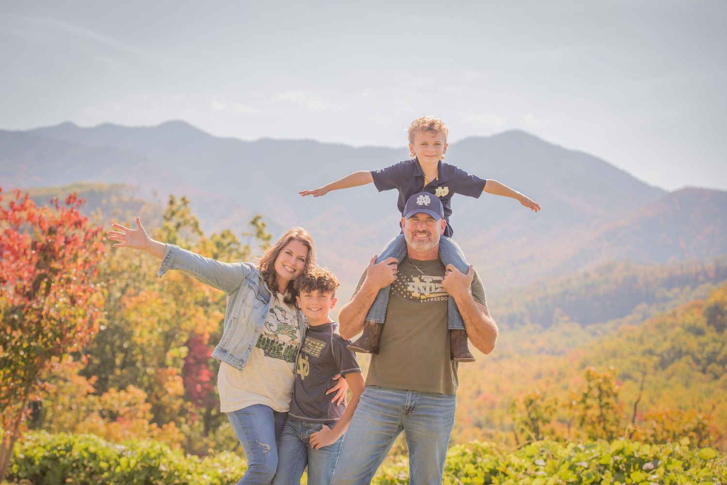 A happy group poses with outstretched arms against a backdrop of autumn mountains and colorful fall foliage.