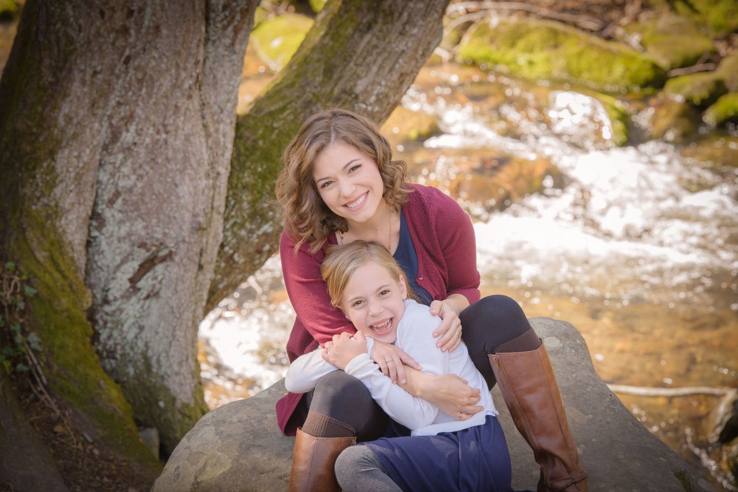 Mother and child people sit together smiling by a tree trunk near a flowing stream on a sunny autumn day Gatlinburg TN.