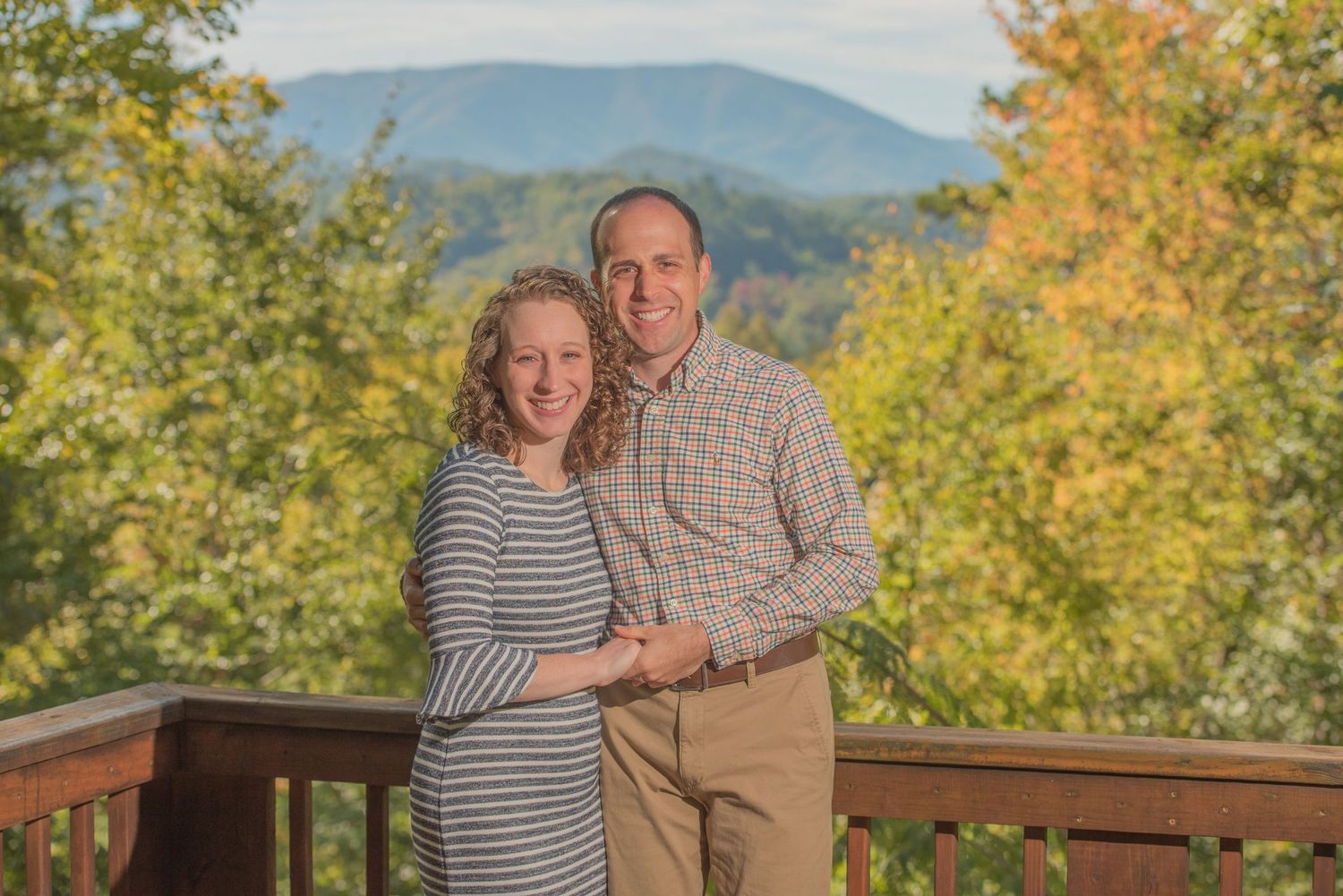 A couple stands on a wooden deck overlooking mountain views with fall foliage in the background. with professional lighting in Sevierville.