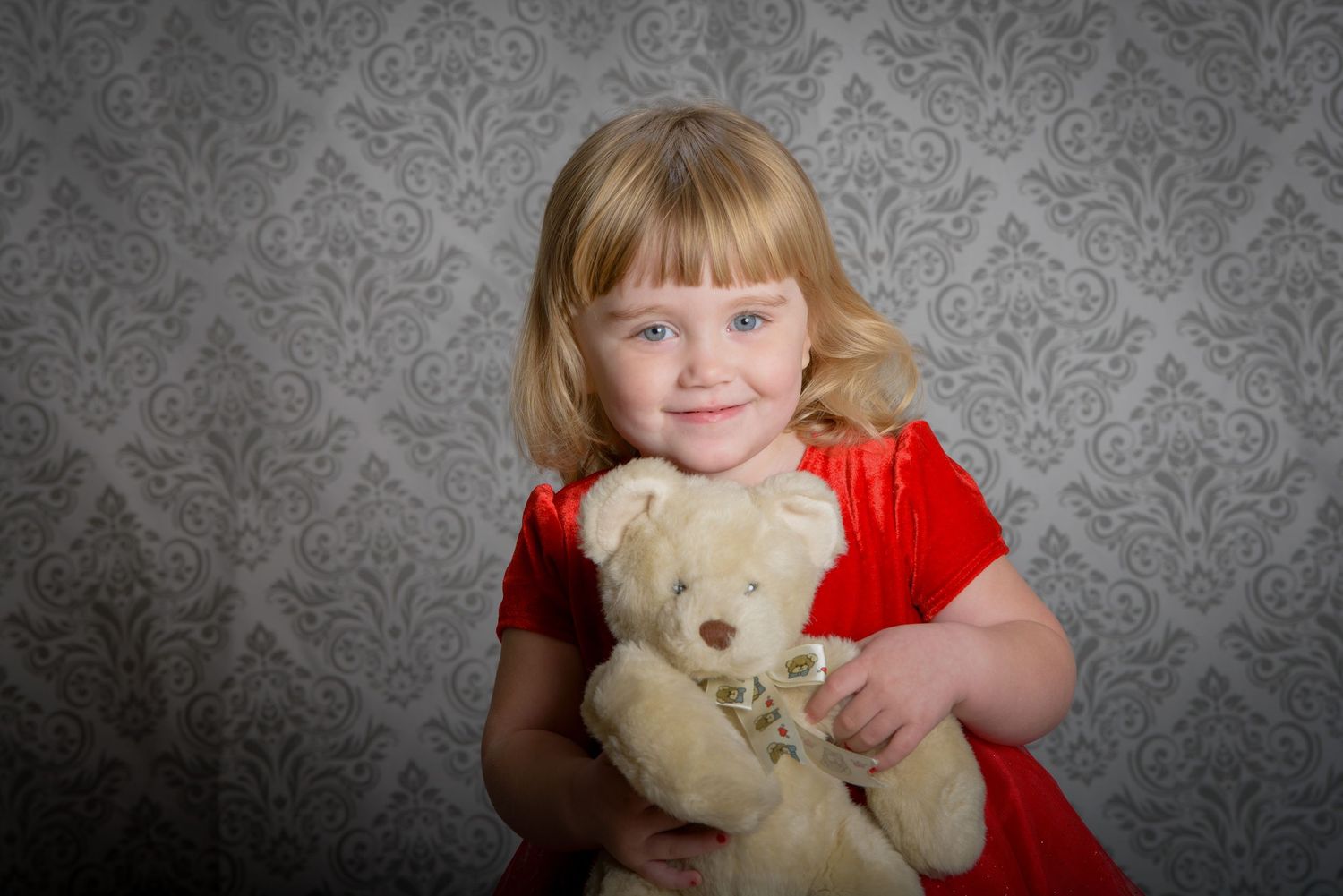 A young child in a red dress smiles while holding a light-colored teddy bear against a gray damask wallpaper background.