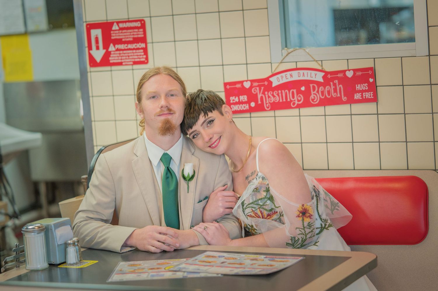 A couple sits together at a diner booth with red seats against a tiled wall, dressed in formal attire and a green tie by Gatlinburg Weddings