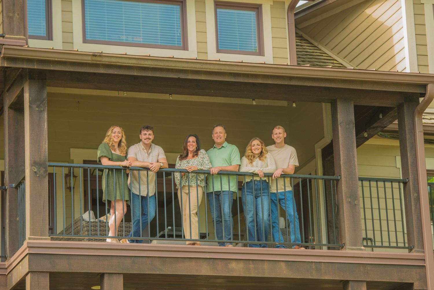 A group of people stand together on a covered balcony of a green house with wooden railings at the rental by the lake in Dandridge TN.