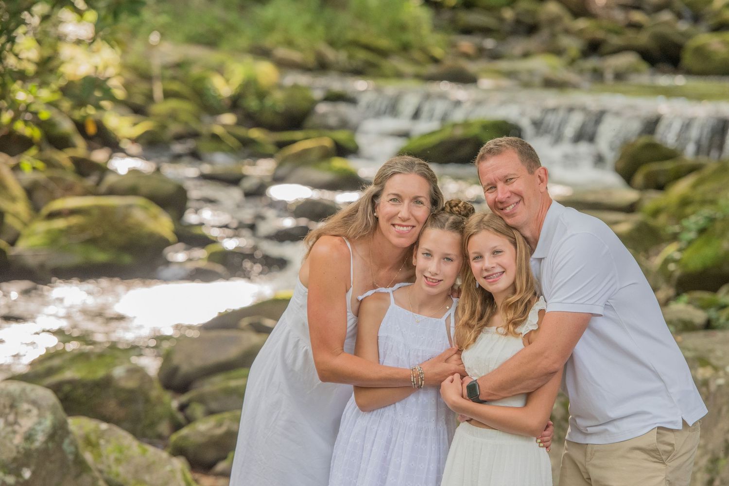 Family in white attire pose together for a portrait by a scenic waterfall with mossy rocks and flowing stream in Townsend TN.