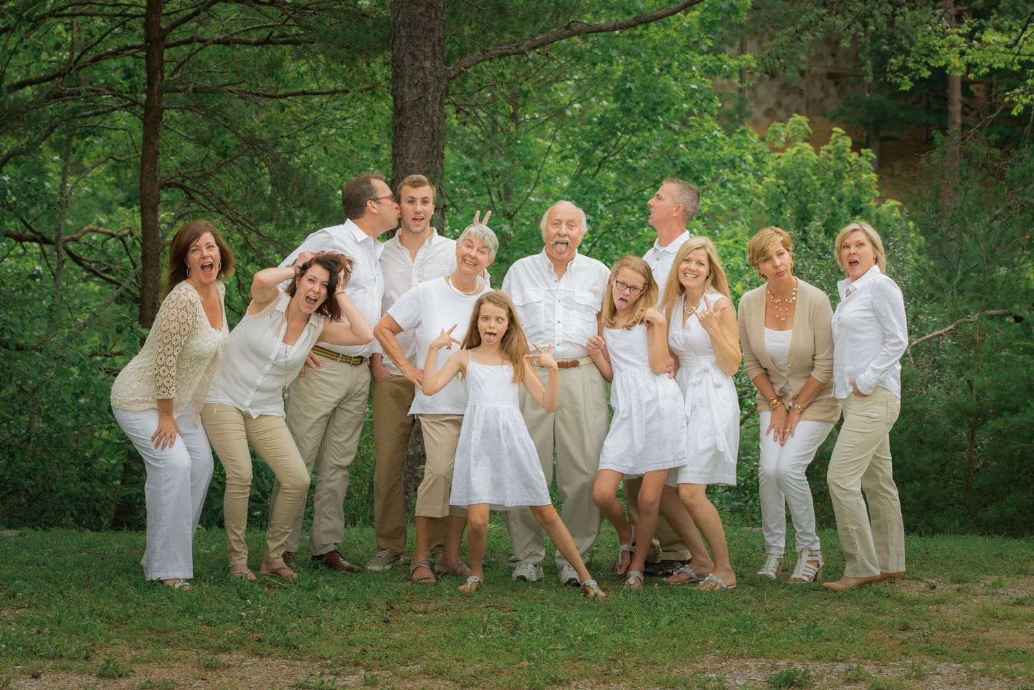 A group in coordinated white and beige outfits poses together on a grassy area with lush green trees in the background.