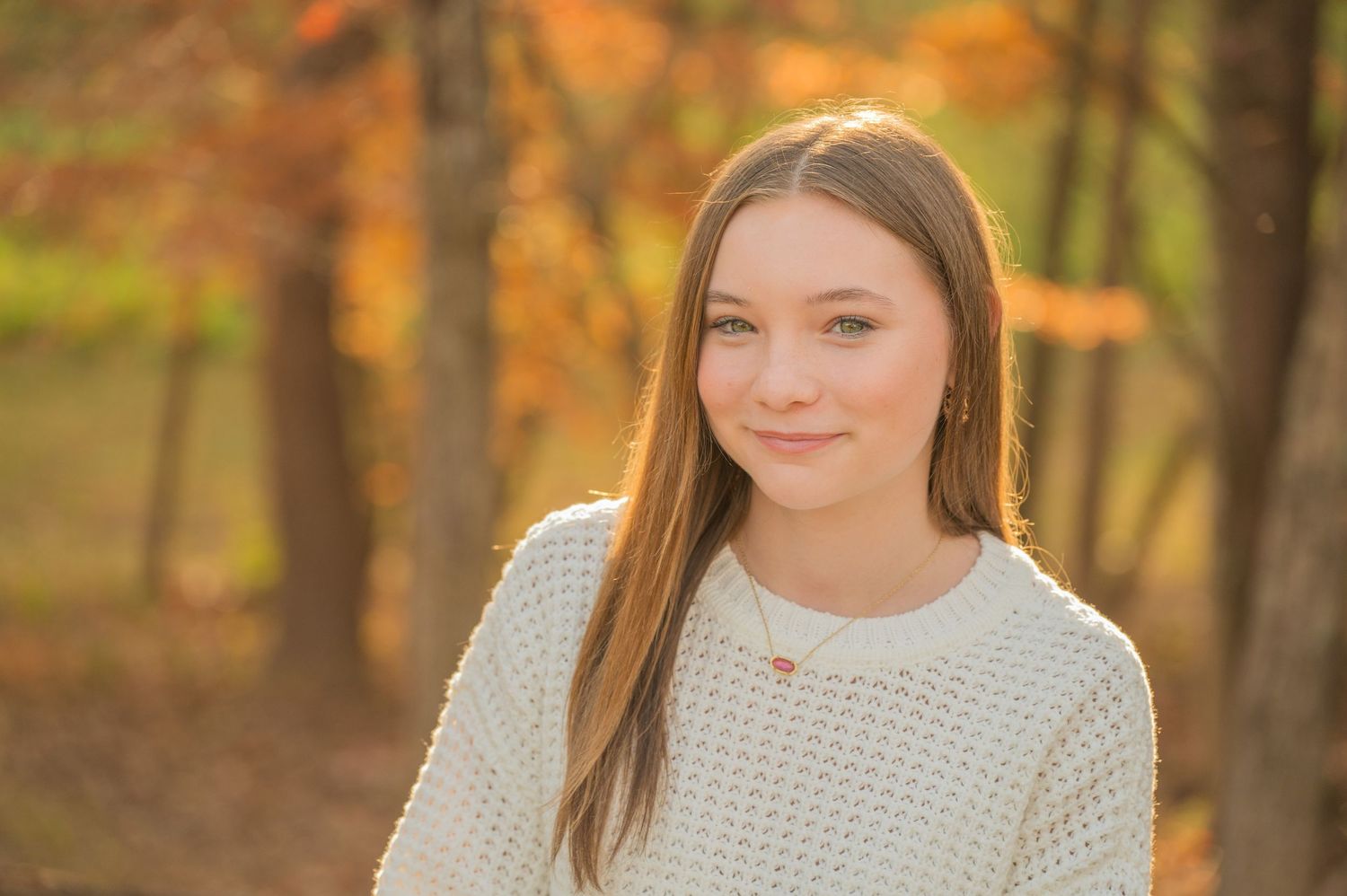 A girl in a cream knit sweater smiles warmly in a forest setting with autumn foliage in the background in Gatlinburg TN.