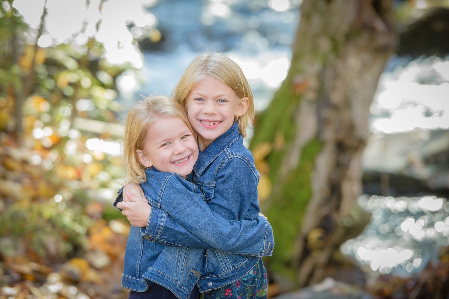 Two sisters in denim jackets embrace and smile while standing near water with autumn foliage in the background in Sevierville TN.