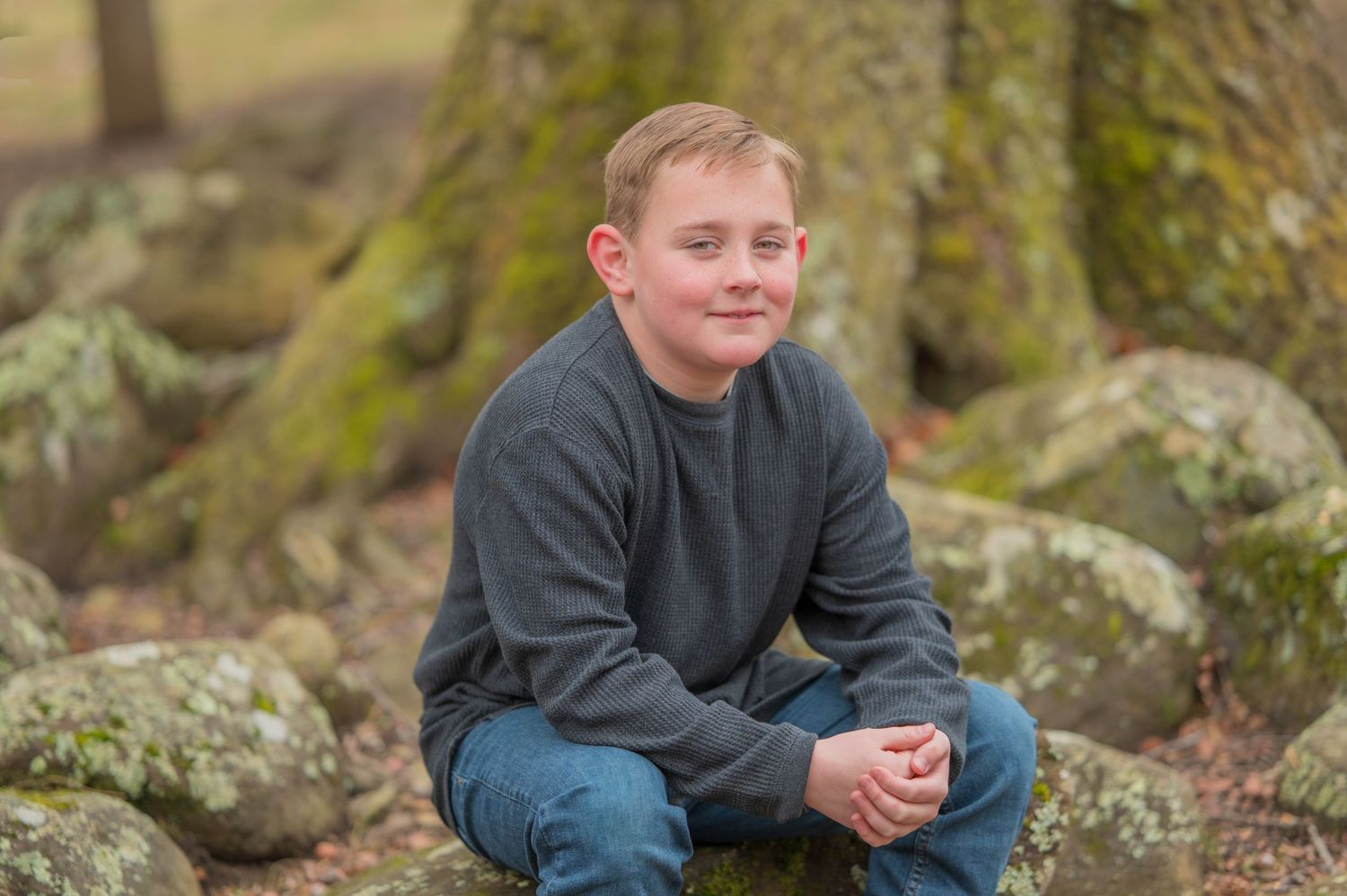 Child in gray sweater and jeans sits casually on mossy rocks in an outdoor nature setting in Sevierville TN.