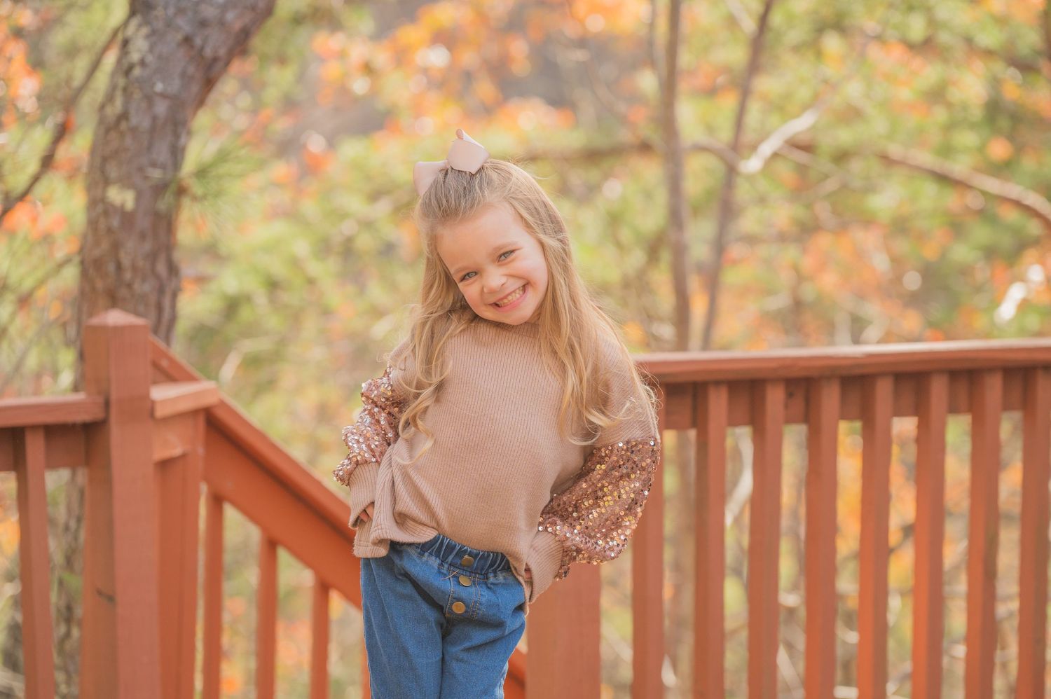 A young child wearing a beige sweater and jeans stands on a wooden deck surrounded by autumn foliage in Pigeon Forge TN.