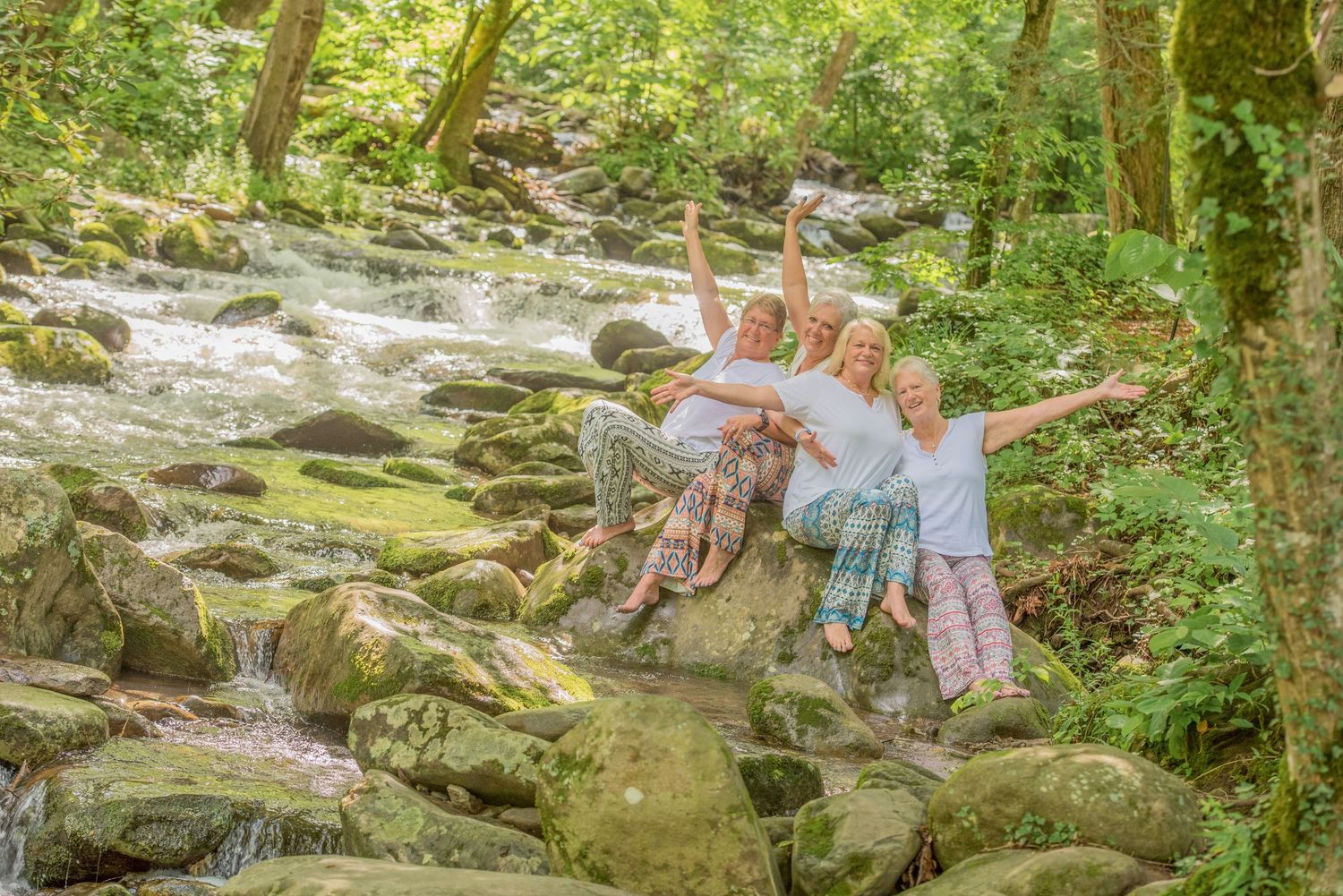 A group of sisters relaxes together on rocks beside a flowing stream in a lush green forest setting by Gatlinburg Photographer TN.