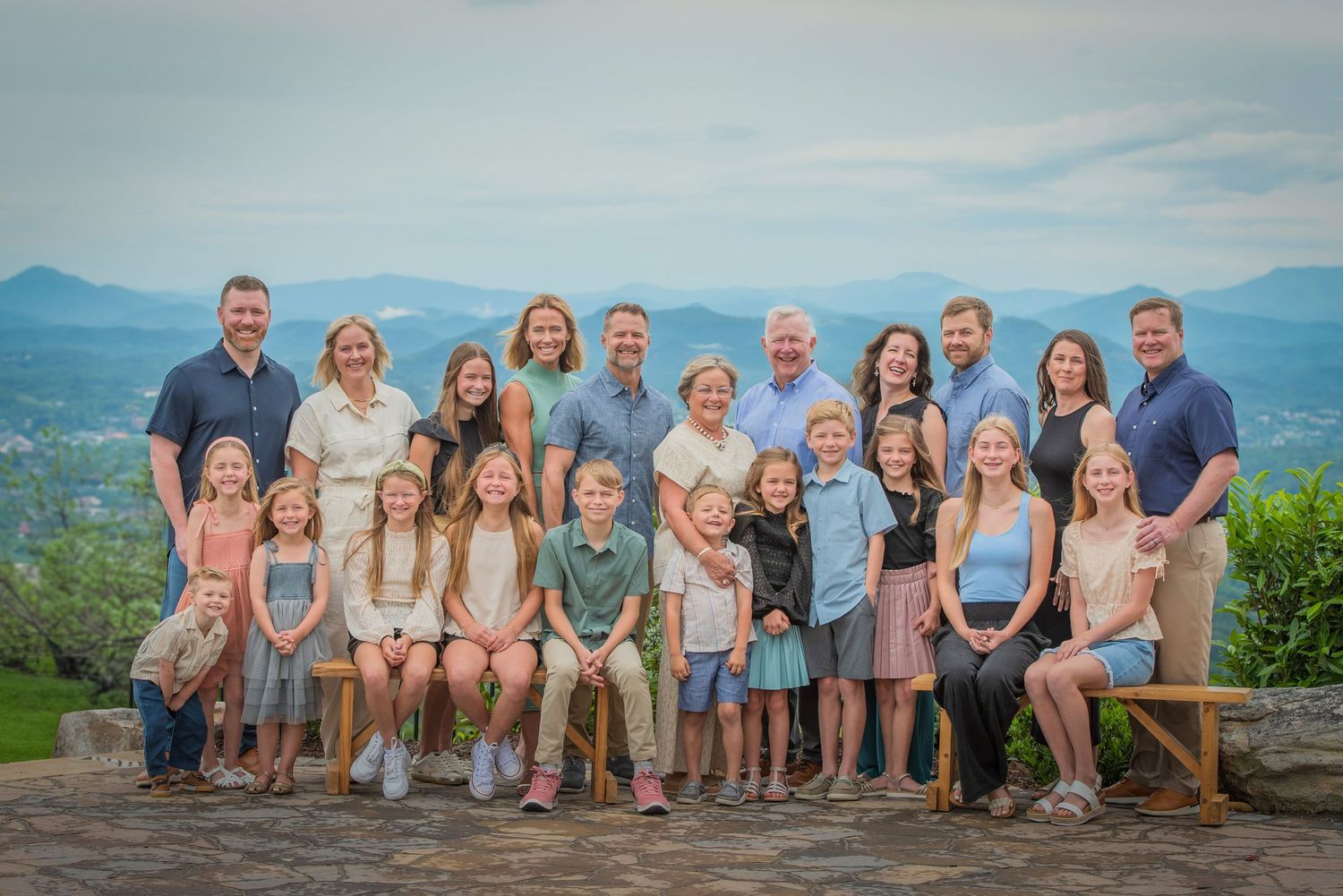 A large family reunion poses together on a mountain overlook with scenic blue ridges visible in the background at their cabin rental.