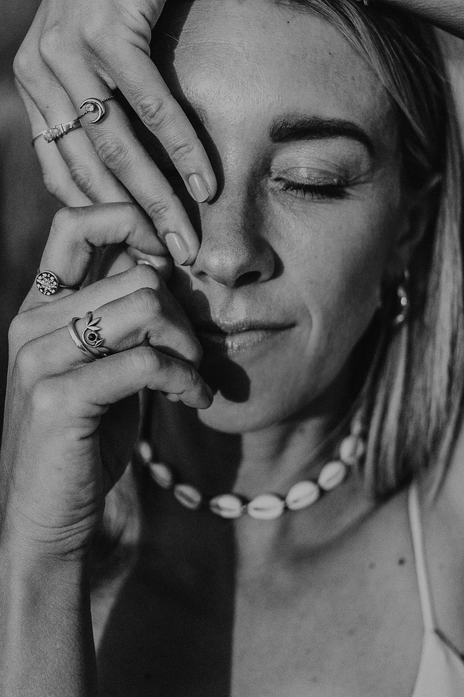 Black and white portrait showing a closed-eyed pose with decorative rings and a shell necklace in an artistic composition.