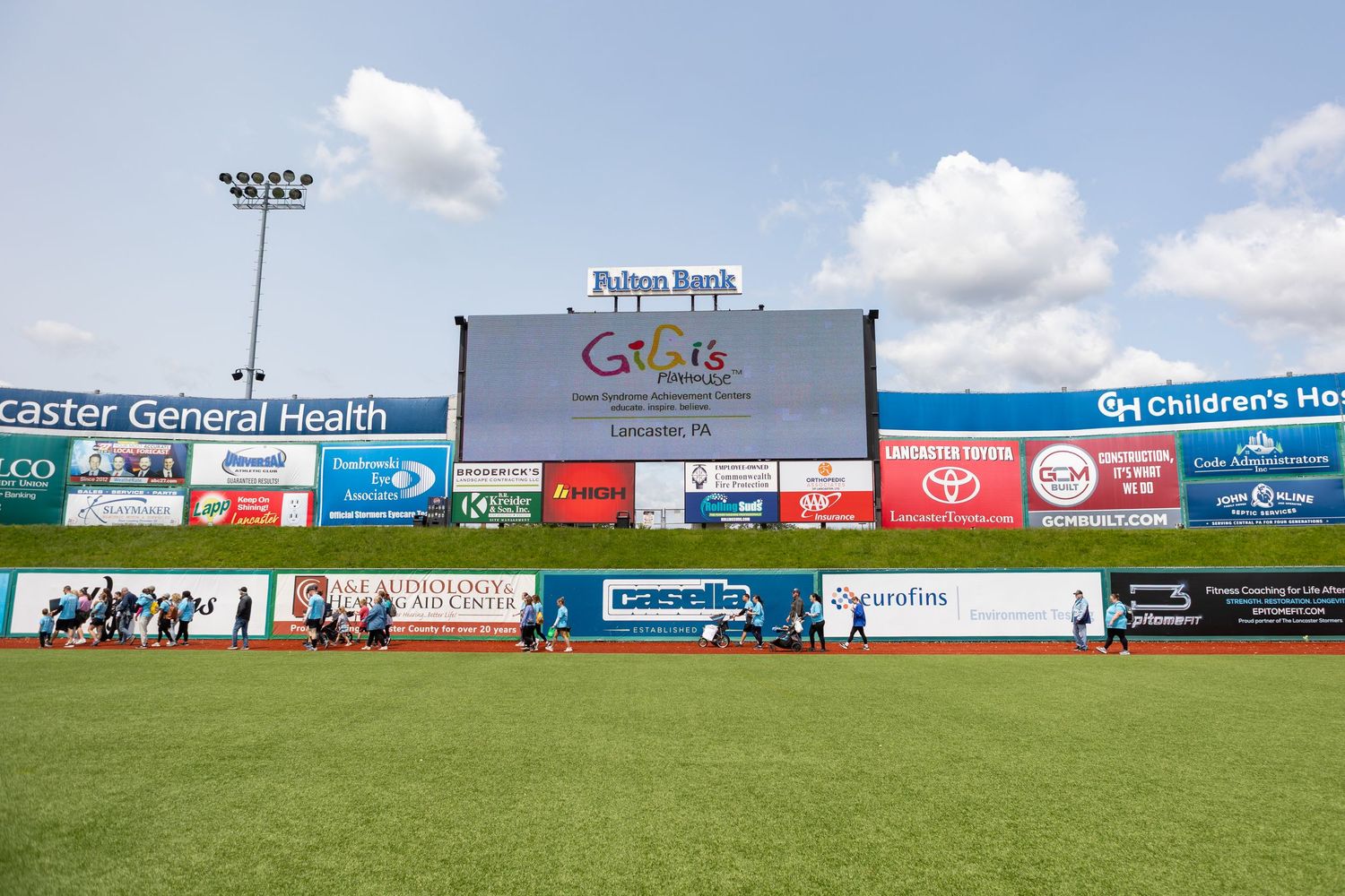 Sports stadium outfield with sponsor signage and advertisements visible along the back wall on a sunny day.