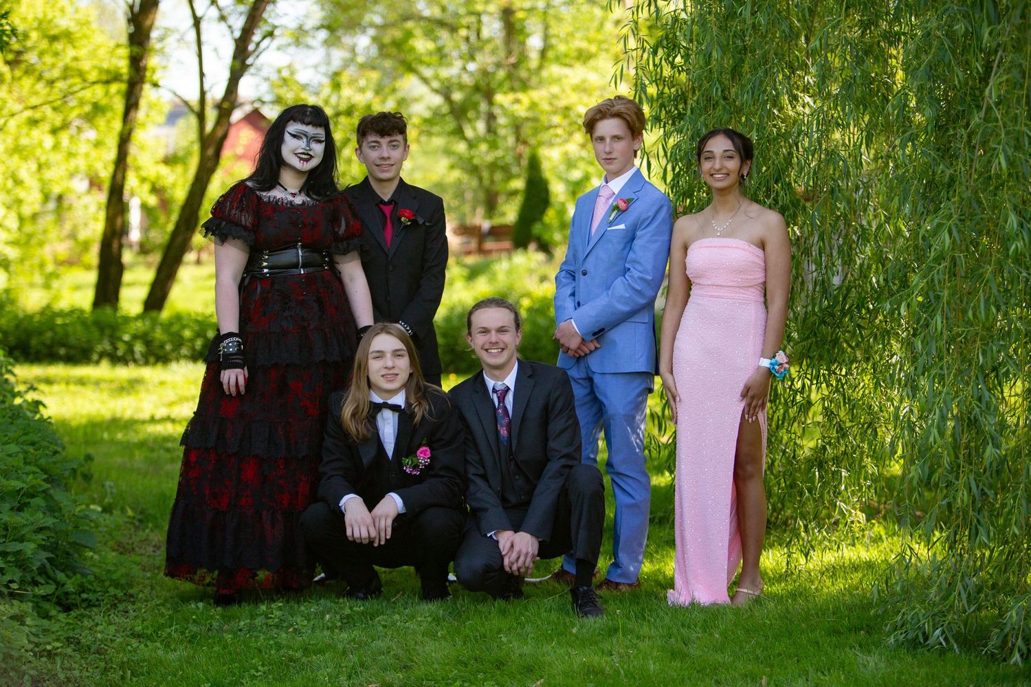 A group poses together outdoors in formal attire under green willow trees on a sunny day.