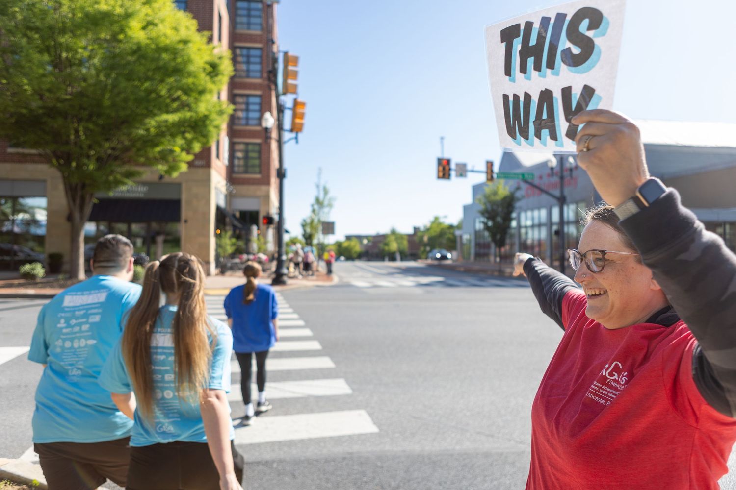 Person holds directional sign while participants in blue shirts cross street during community event.