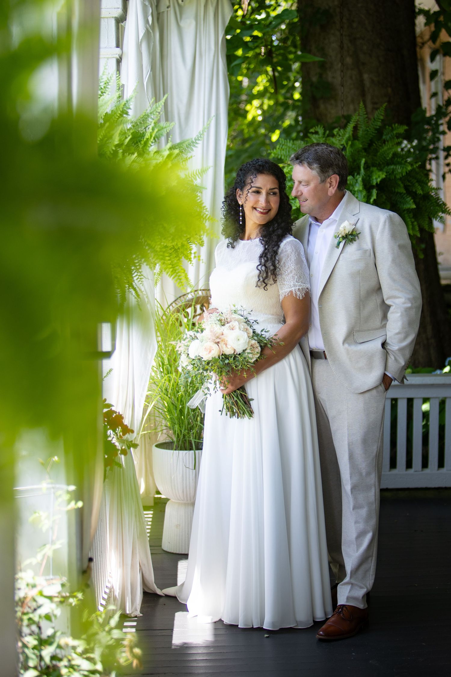 A wedding couple shares a tender moment on a porch surrounded by greenery and white flowers.