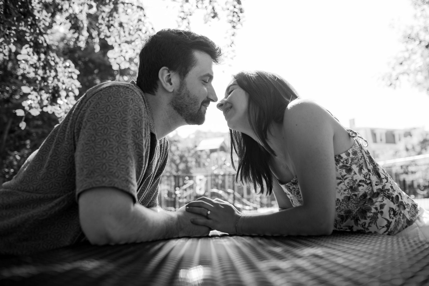 Black and white photo of a couple lying on a picnic blanket leaning in for a romantic kiss outdoors.