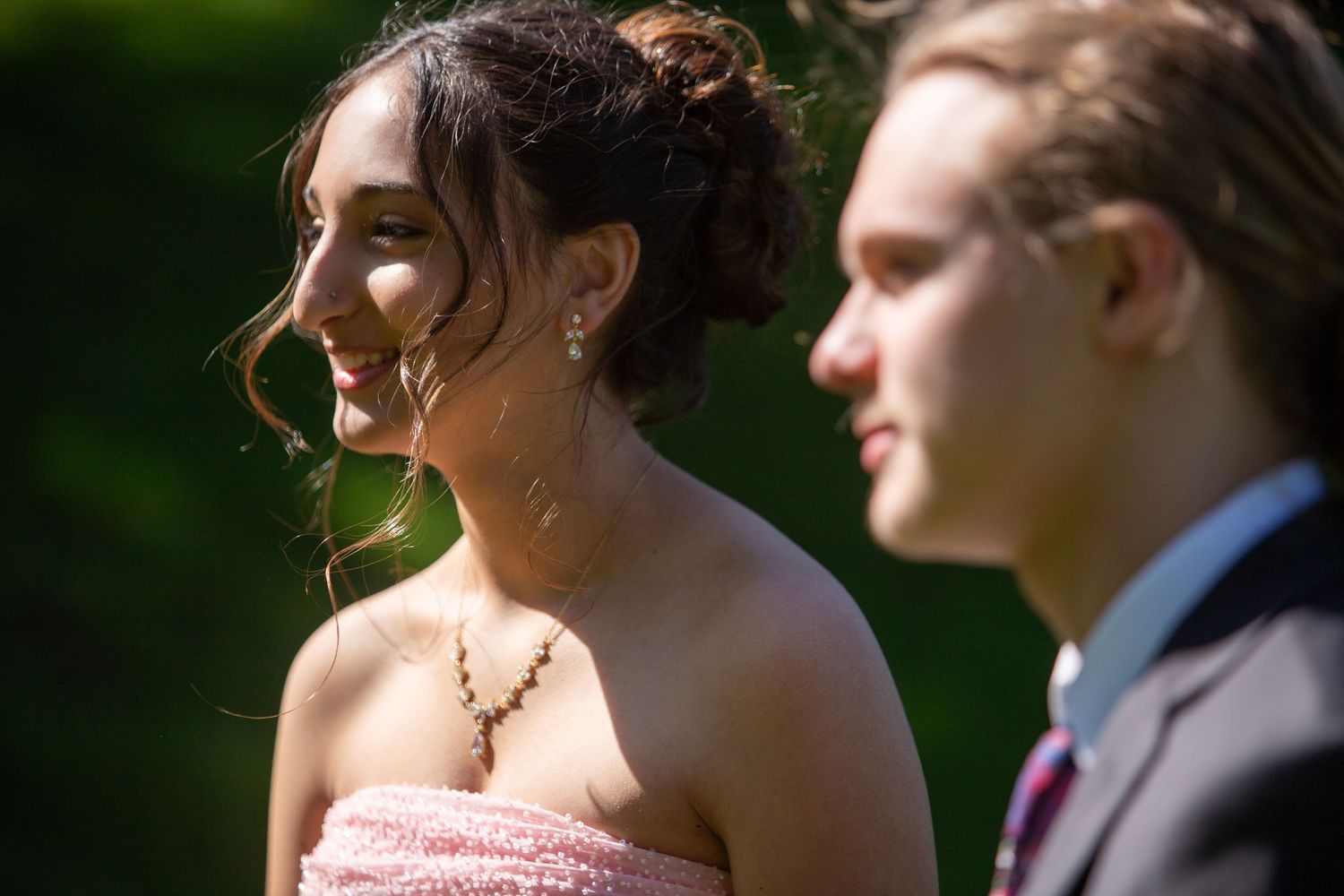 A couple dressed in formal attire stand together outdoors with soft sunlight illuminating them against a green background.