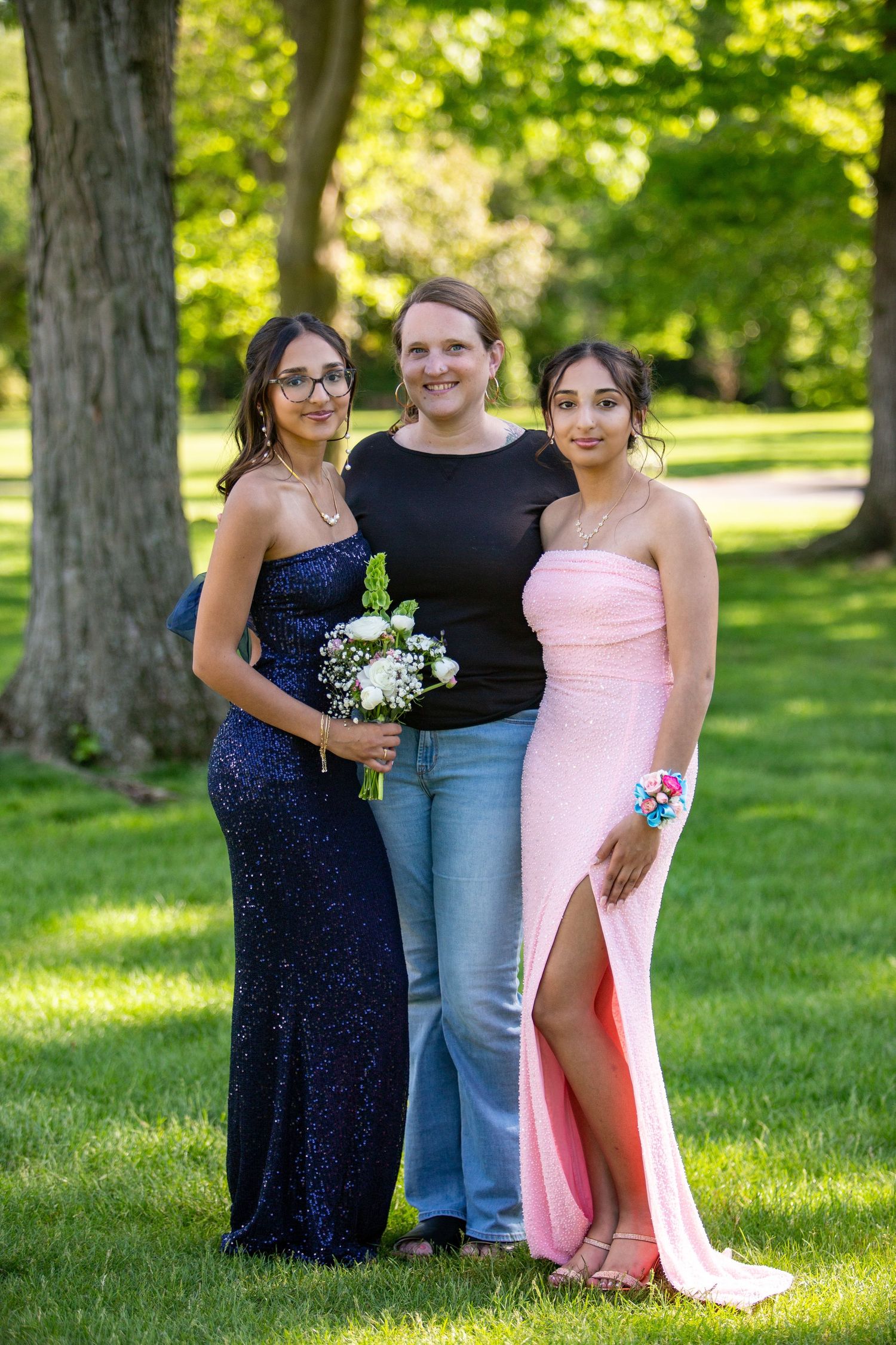 Three people in formal attire stand together on a sunny lawn with white flowers, wearing navy blue and pink dresses.