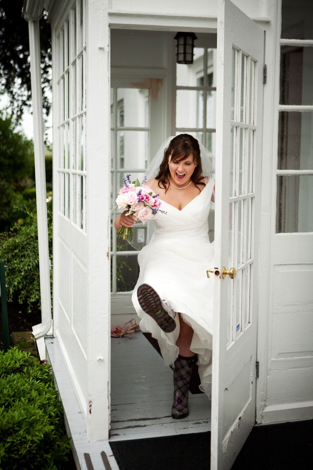 A bride wearing boots stands playfully in a white doorway while holding a pink and white flower bouquet.
