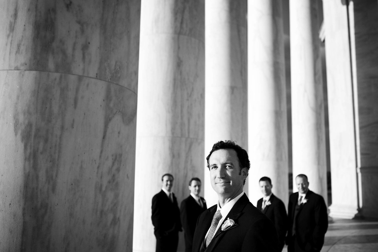 Silhouettes of wedding party members stand between dramatic white columns in black and white photography.