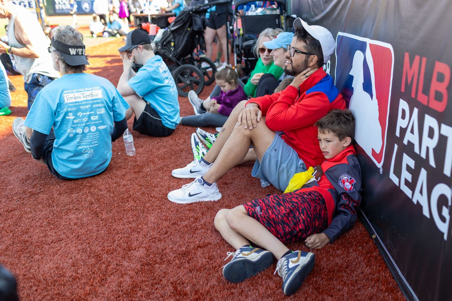 Group of participants sitting along the baseball field warning track during an outdoor event.