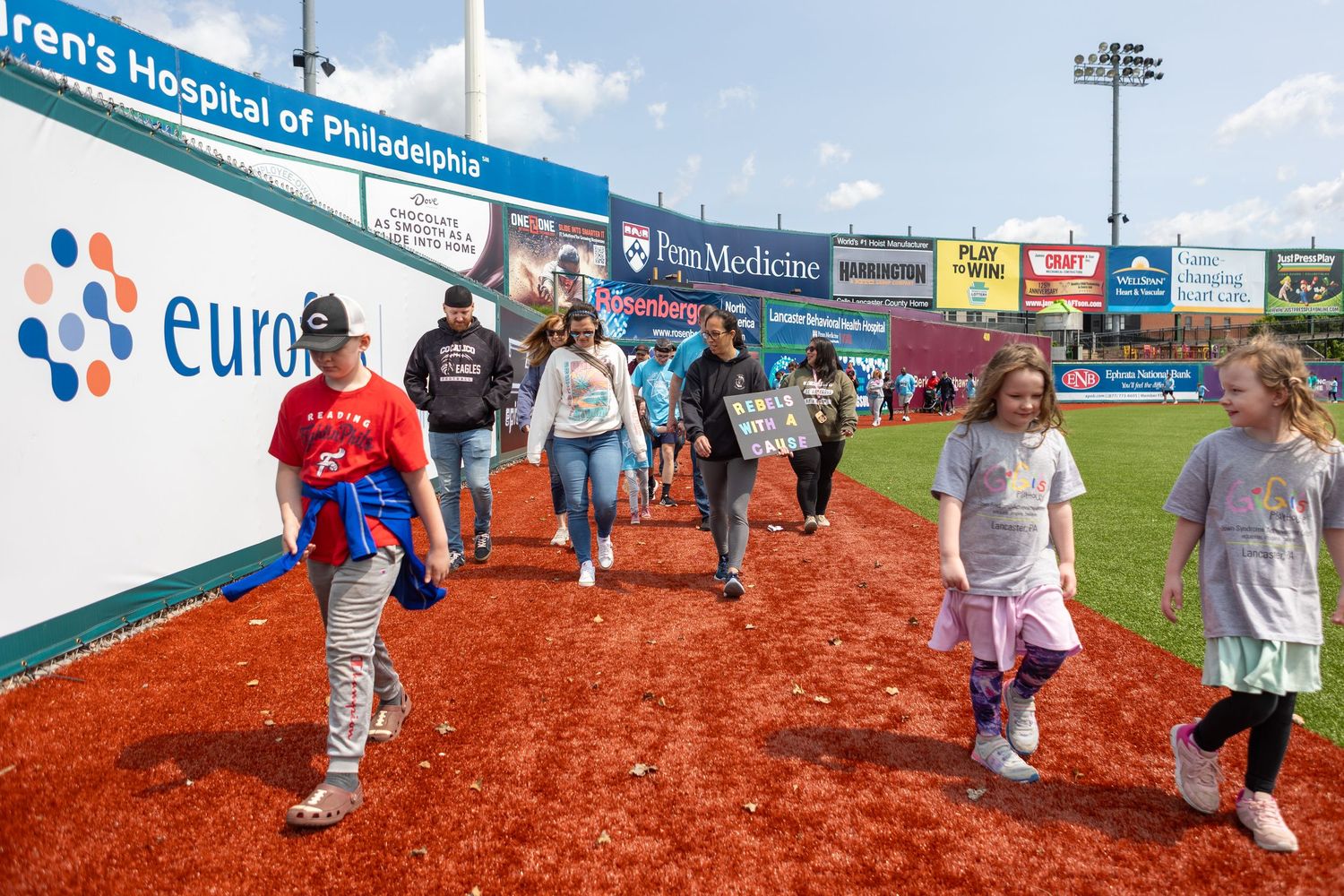 Children and adults walking along a baseball field warning track past stadium advertising banners.