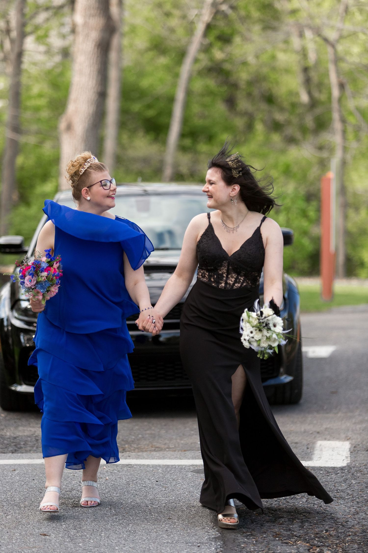 Two women in formal dresses walking hand in hand, one in royal blue and one in black, with flowers and a car in background.