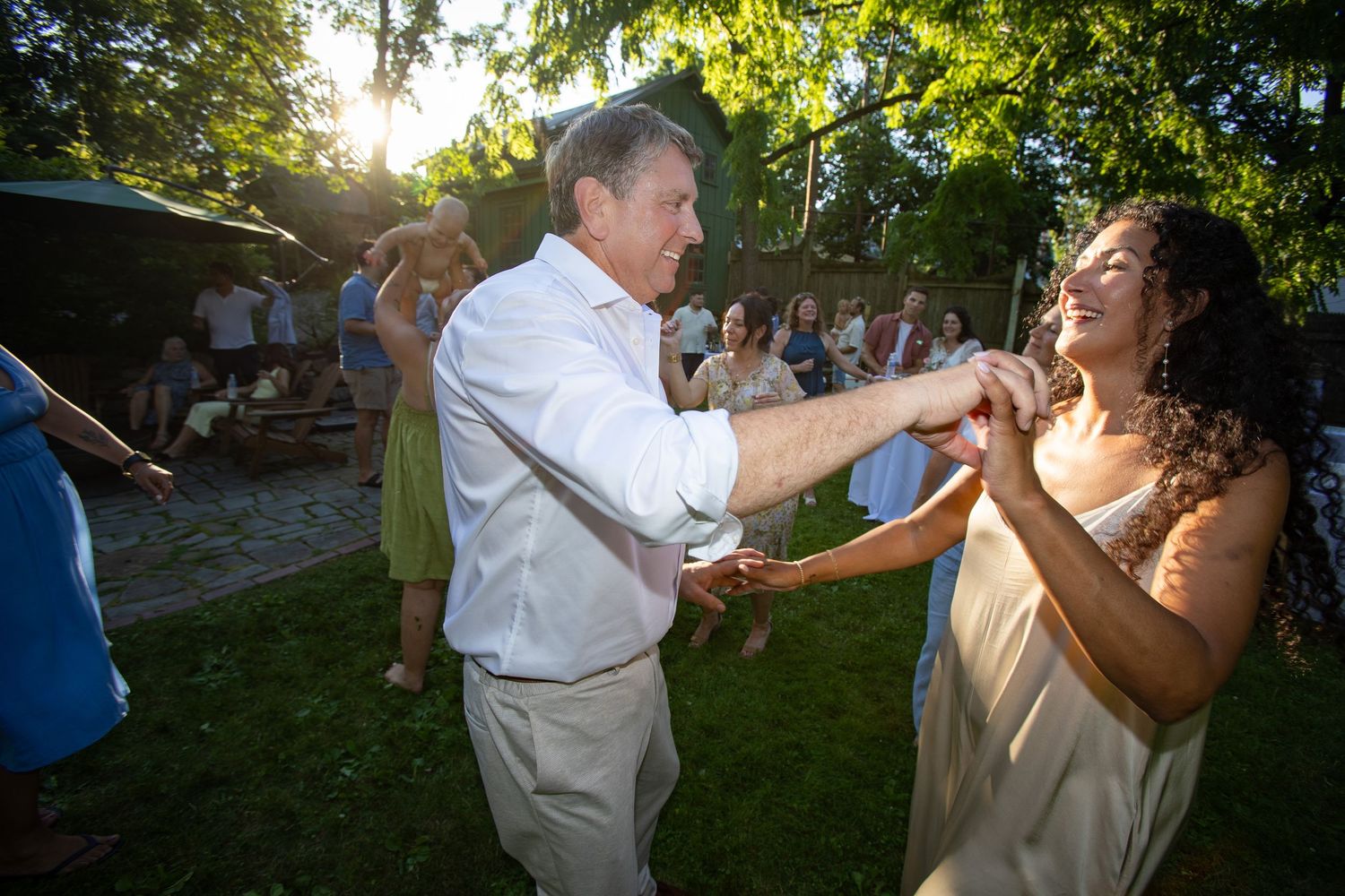 Two people dance together joyfully at an outdoor wedding reception as sunlight filters through trees in the background.
