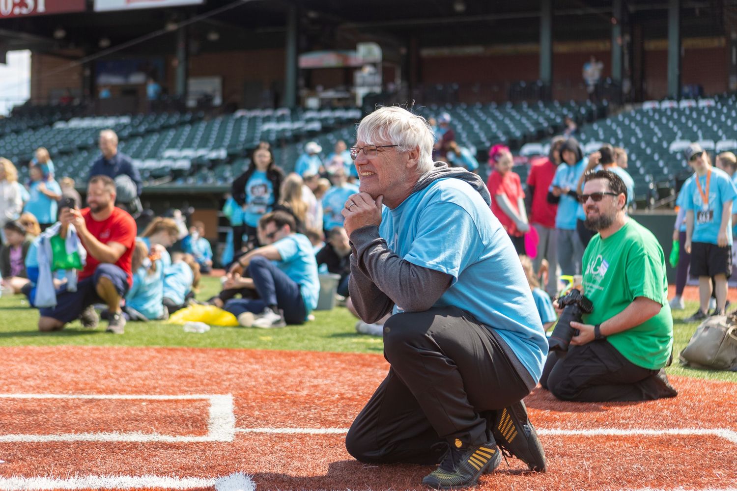 Person crouching at baseball field wearing blue shirt during outdoor event.