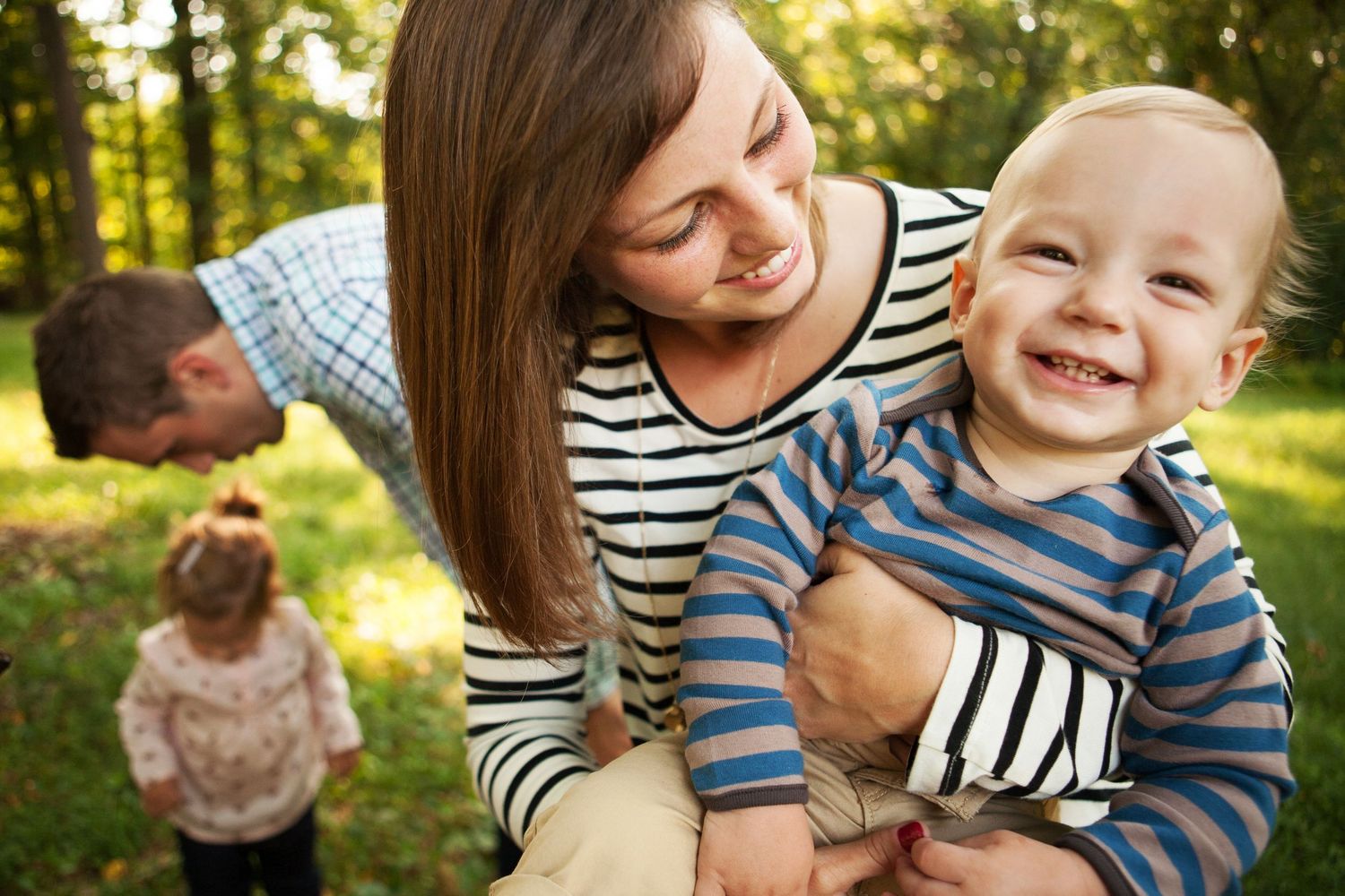 Happy baby in blue striped shirt laughs while being held outdoors in park with sunlit trees in background.