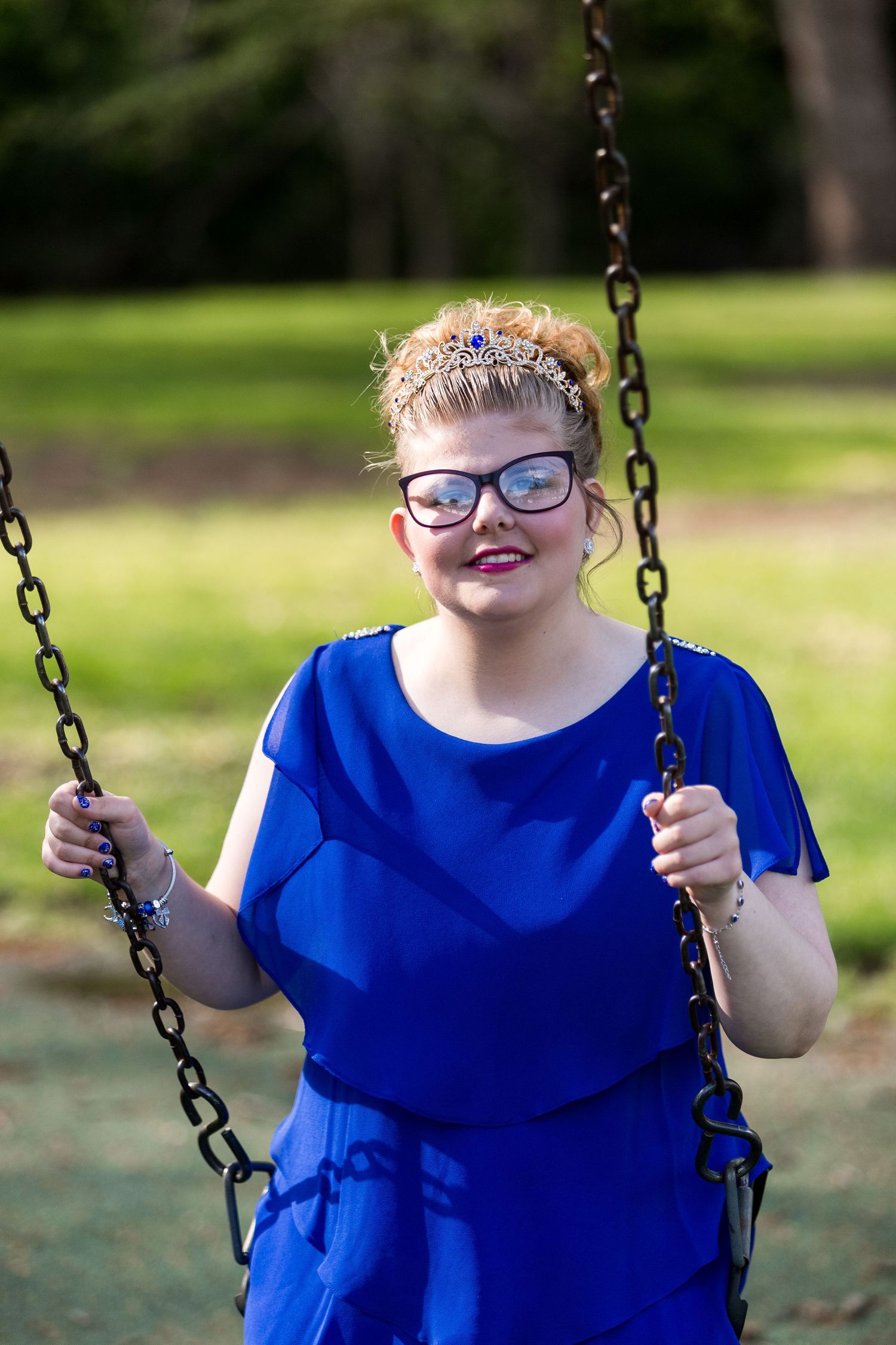 Someone wearing glasses and a bright blue dress sits smiling on a park swing on a sunny day.