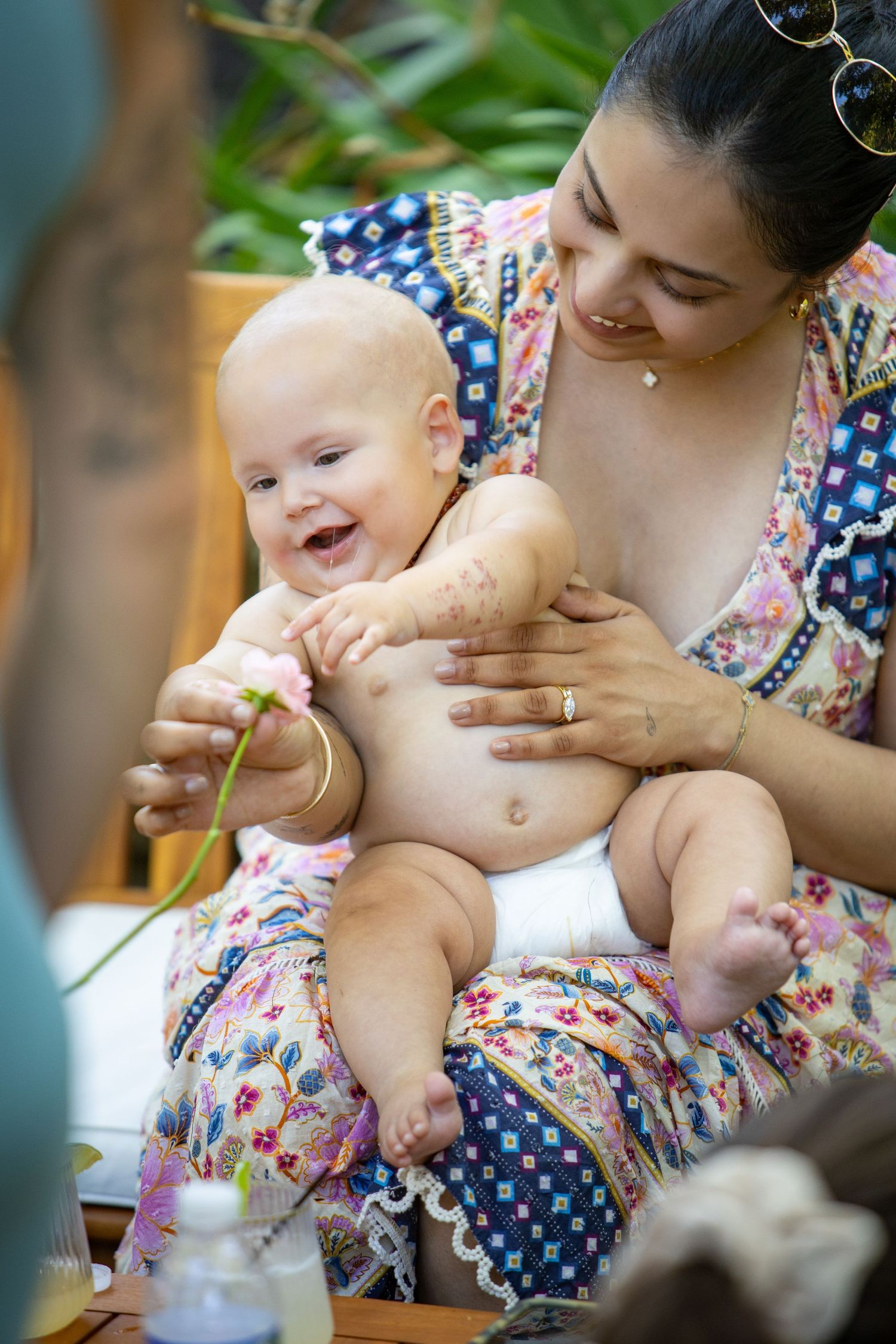 A joyful baby laughs while sitting on someone's lap in a floral dress outdoors.