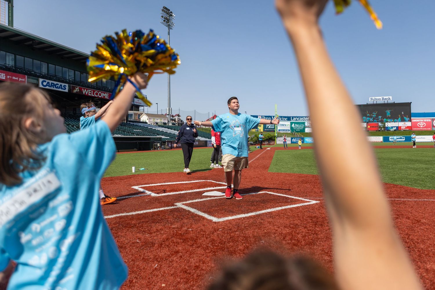 Participants cheer and wave pom-poms on a baseball field during a special event.