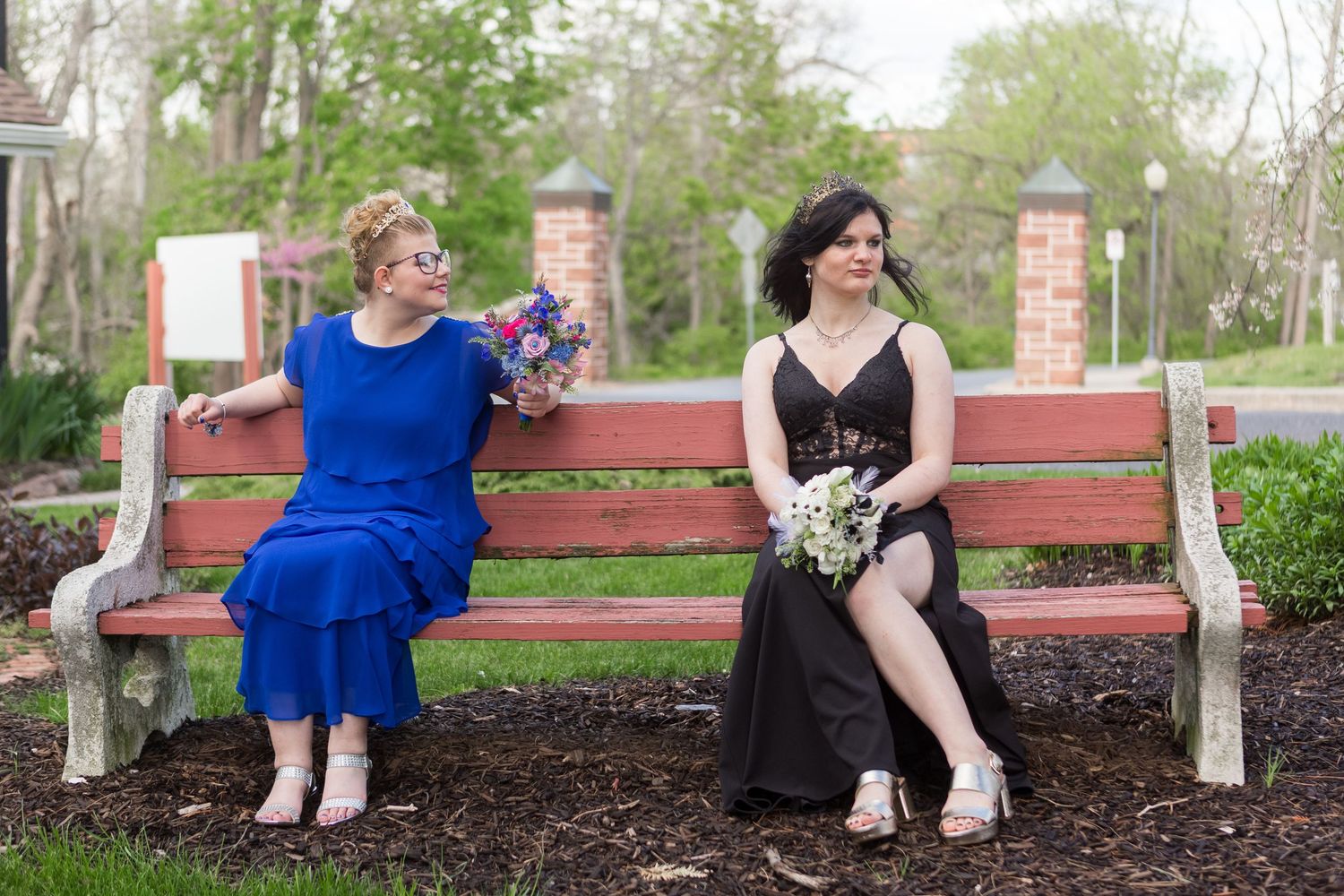 Two elegantly dressed people sit together on a red wooden bench in a garden setting, wearing blue and black dresses.