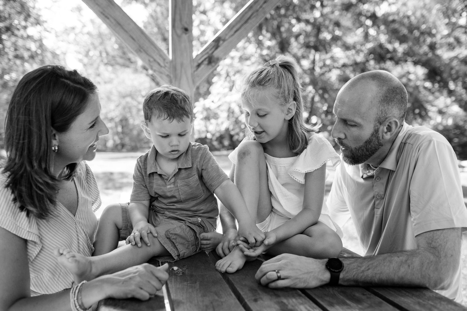 A loving family moment captured in black and white as they gather around a picnic table outdoors on a sunny day.
