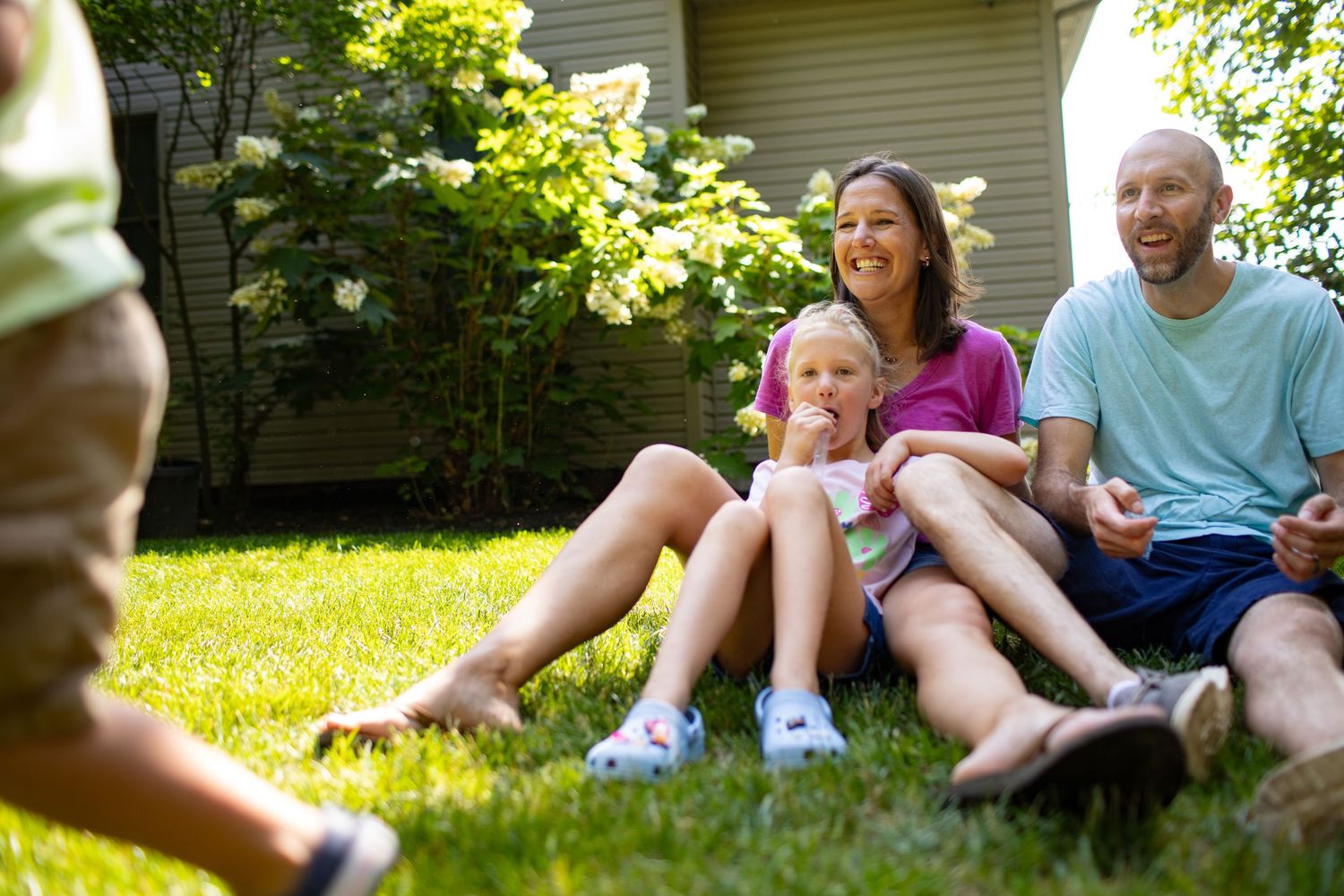 Family sitting together on lush green grass in backyard garden on sunny summer day.