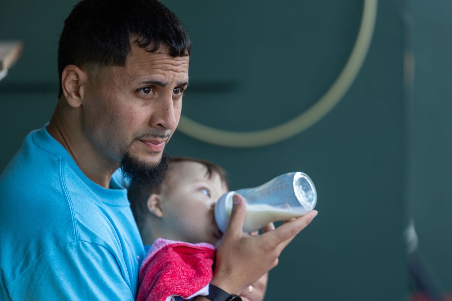Person in blue shirt feeding baby from bottle in stadium setting.