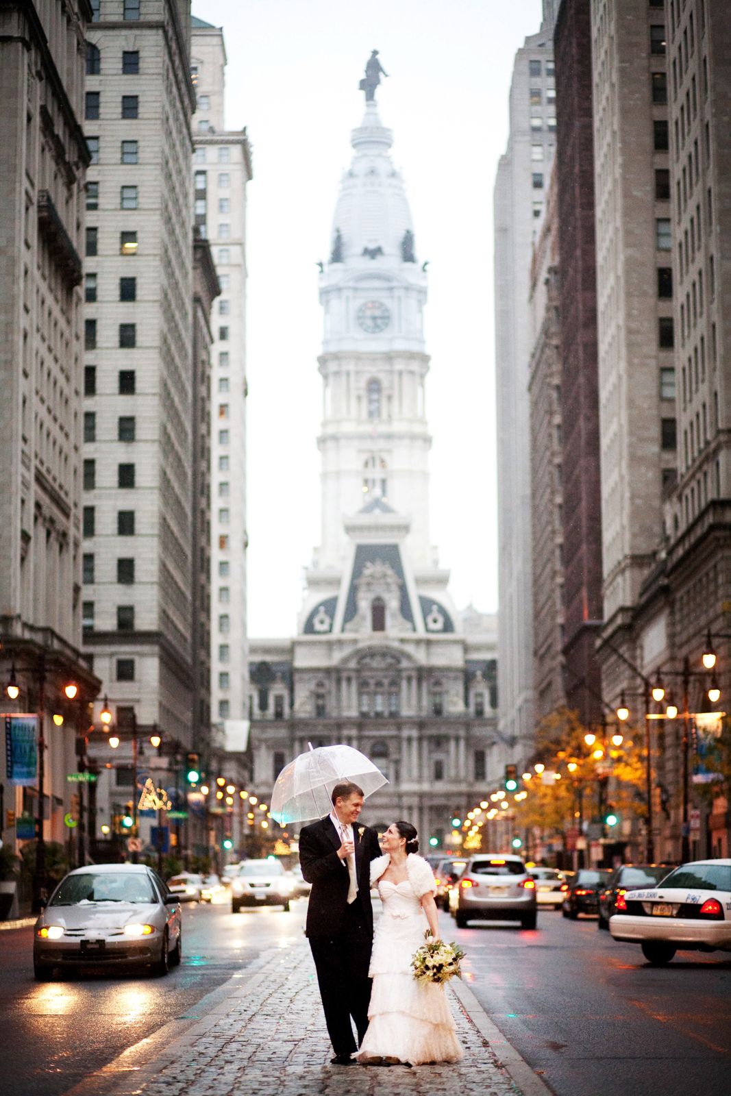 A couple stands on a rainy Philadelphia street with City Hall's historic clock tower rising majestically in the background.