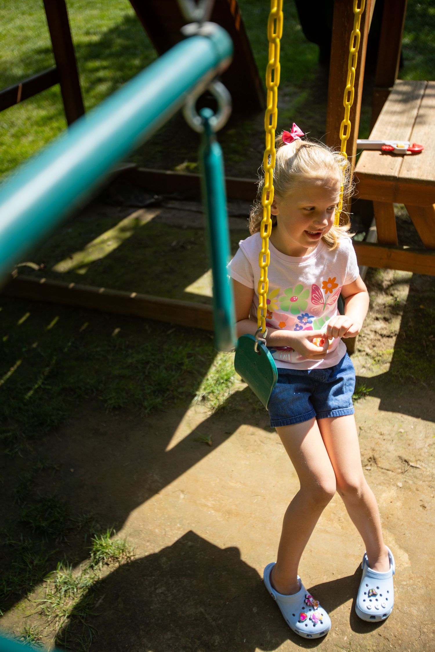Child plays on a wooden playground swing set with blue and yellow equipment in sunny backyard.