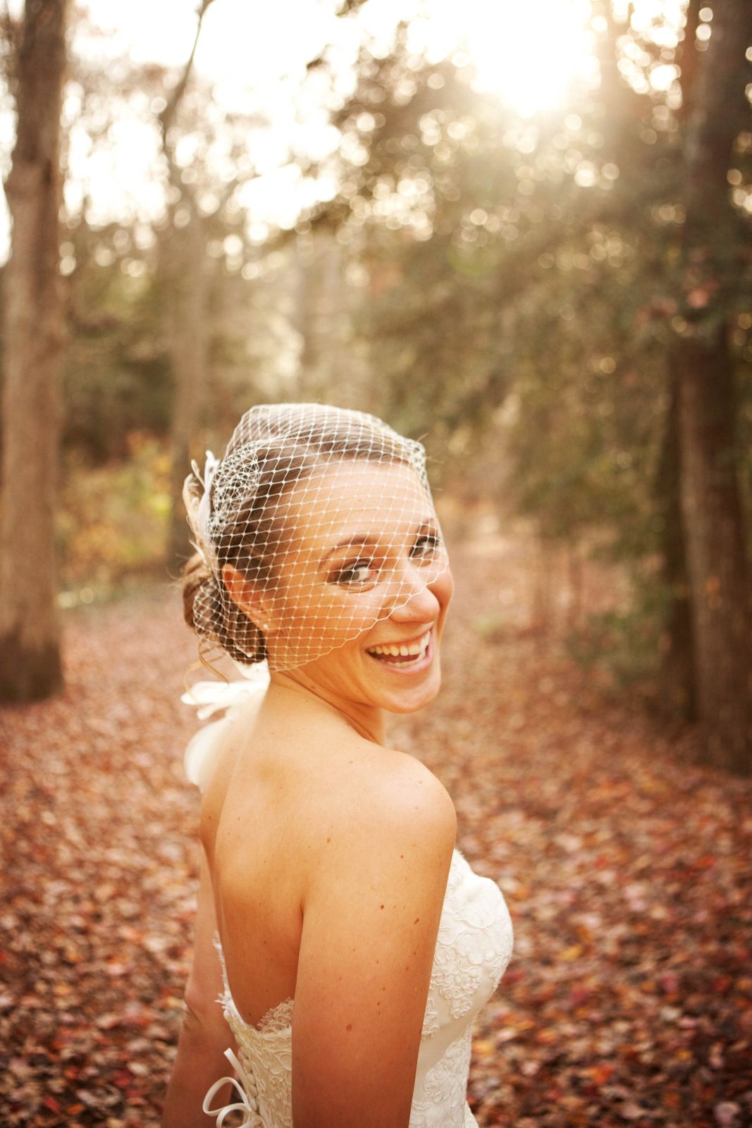 A strapless white wedding dress shimmers in autumn sunlight as golden leaves scatter across a wooded forest path.