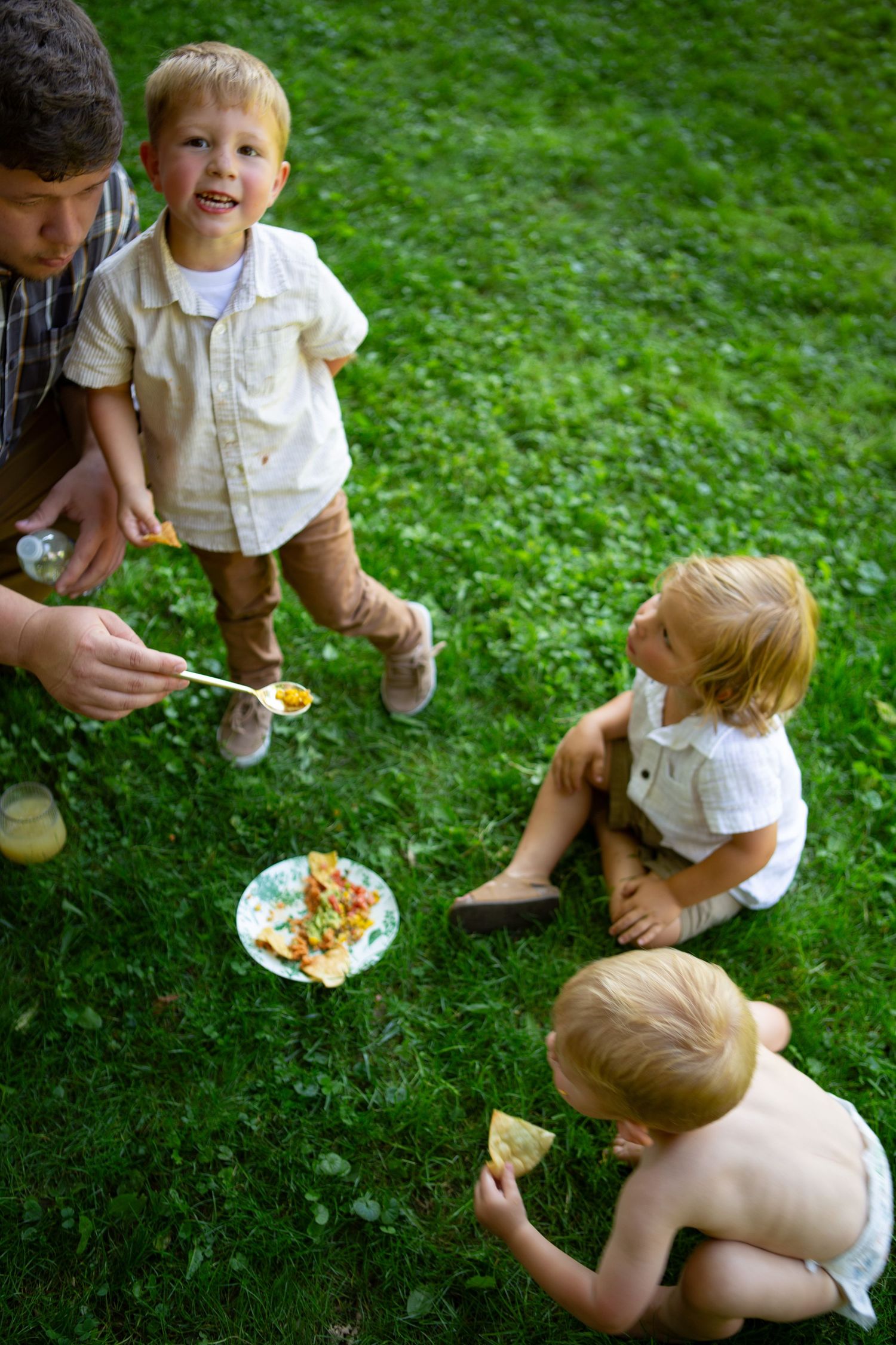 Young children gather on grass while an adult serves food during an outdoor gathering.