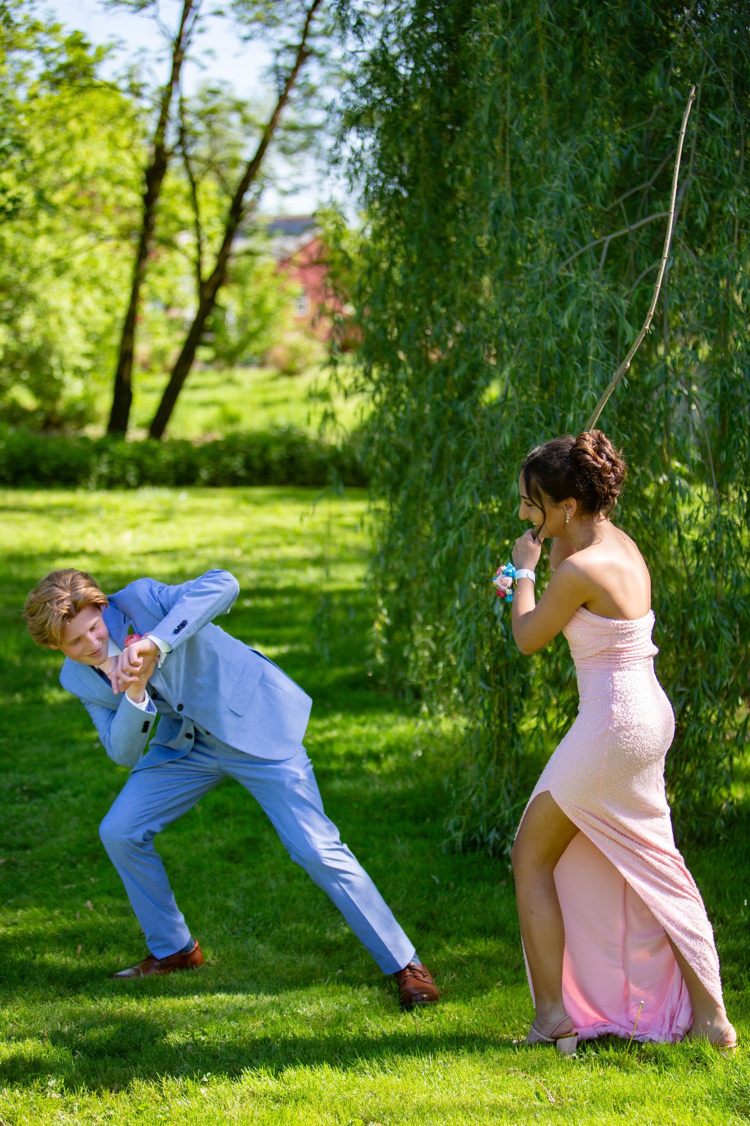 Two people in formal attire playfully strike silly poses while having fun on a lawn under a willow tree.