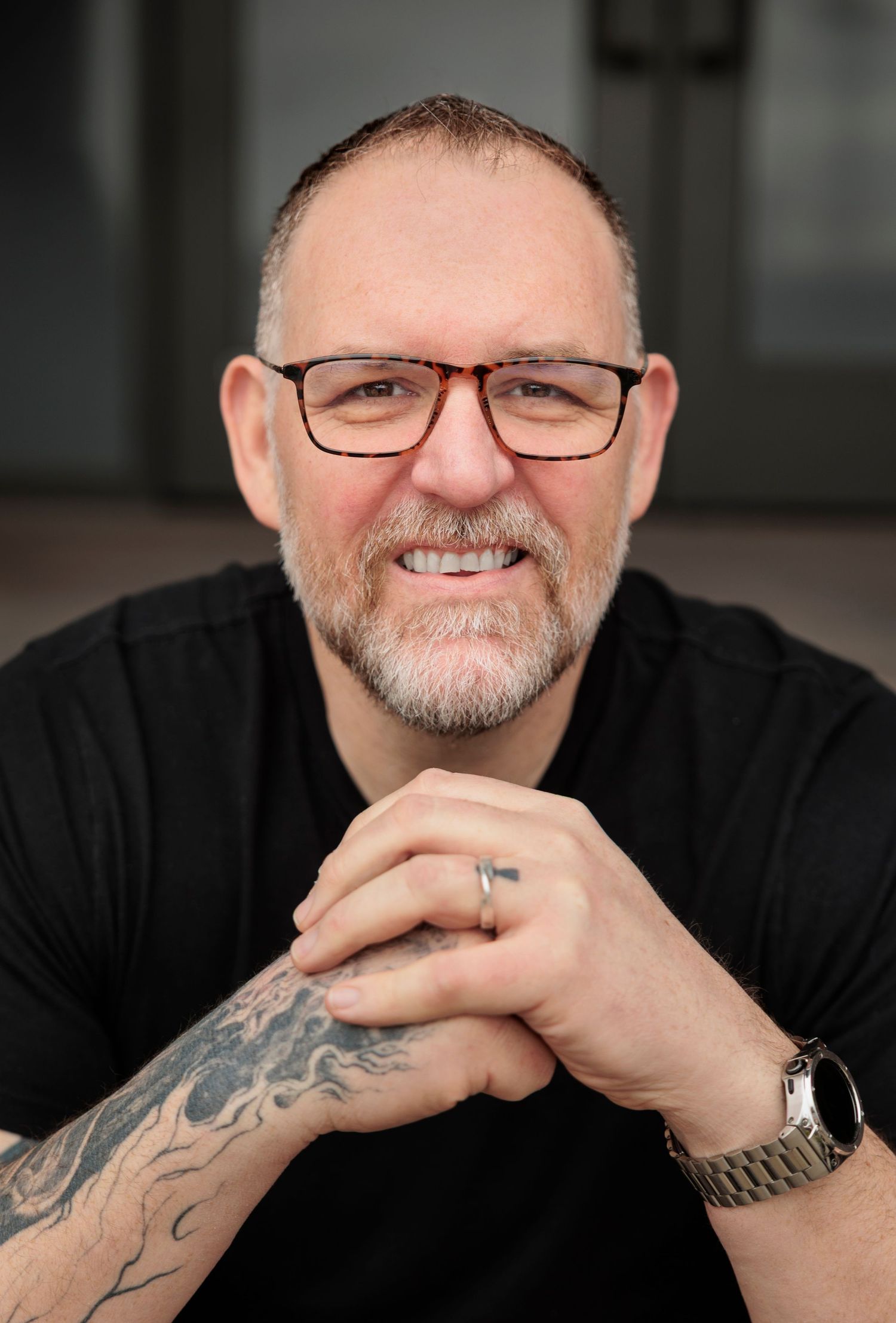 Portrait of person in glasses and black shirt showing tattoos and watch. Joel Wiebner